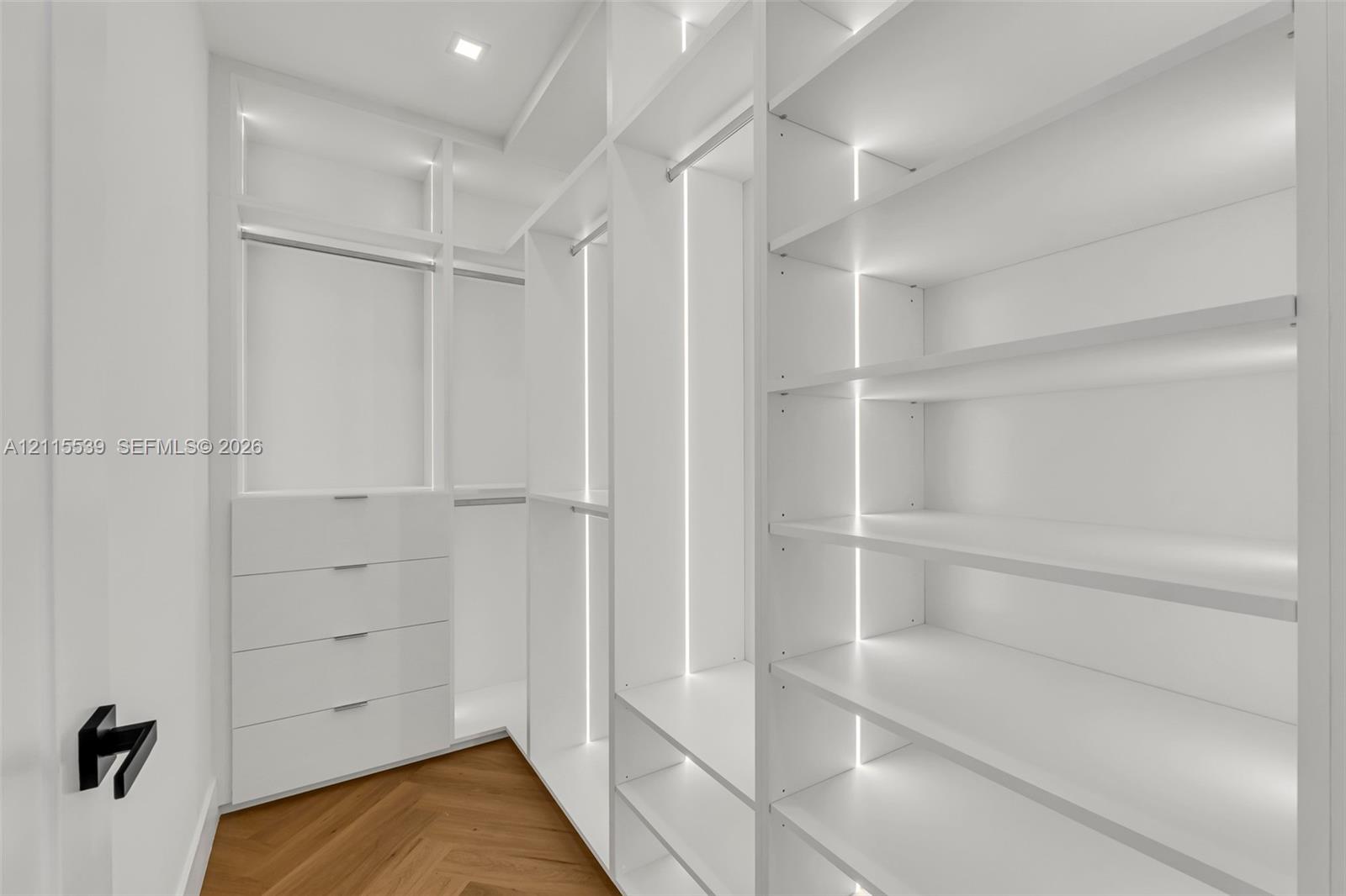 Custom storage system featuring floor-to-ceiling white cabinetry, integrated LED strip lighting, built-in drawer units, adjustable shelving, and herringbone wood-finish flooring