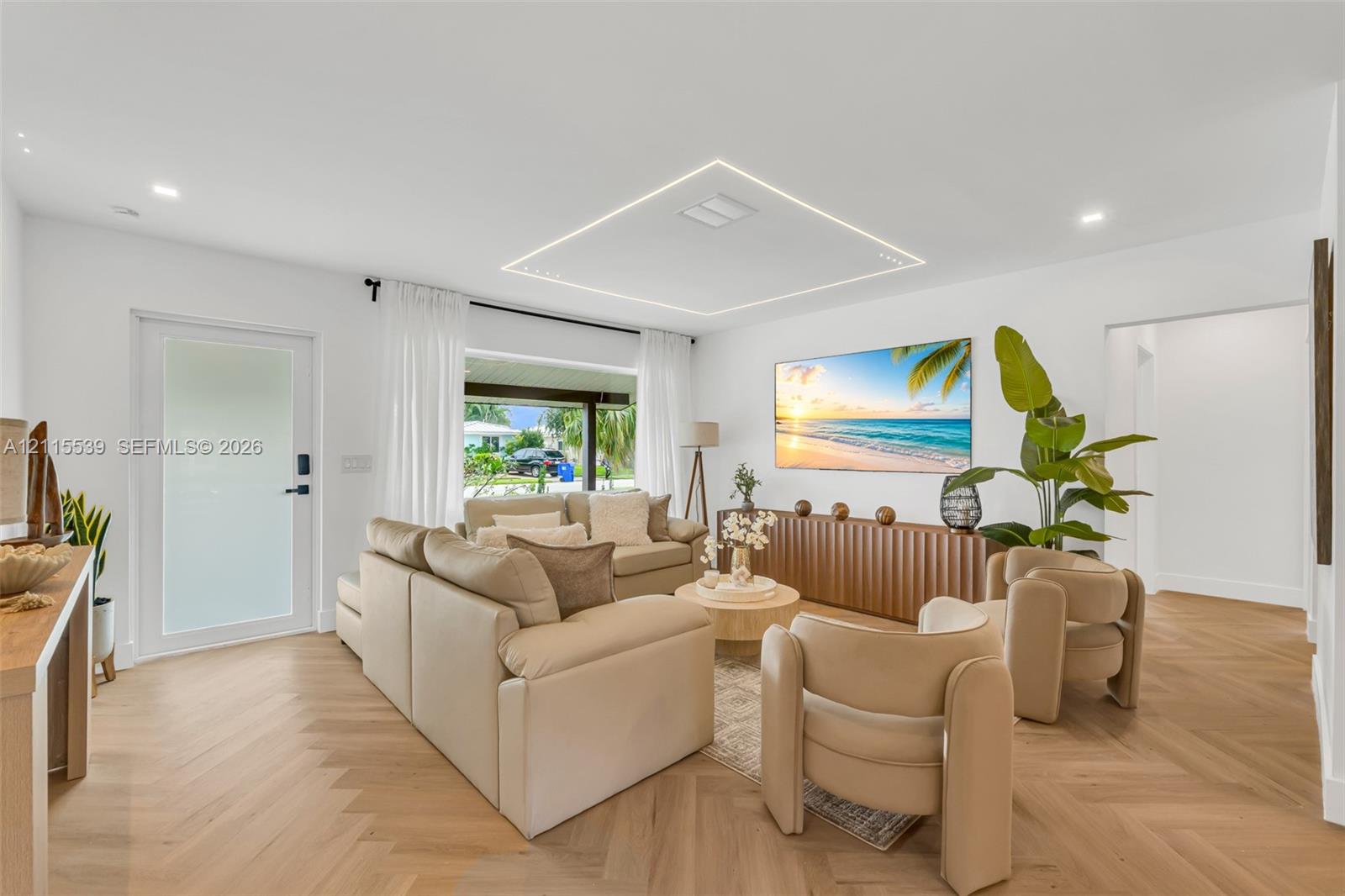Living area featuring light-toned herringbone patterned flooring, recessed ceiling lighting, a recessed rectangular architectural ceiling detail with perimeter illumination, and a frosted glass entry door