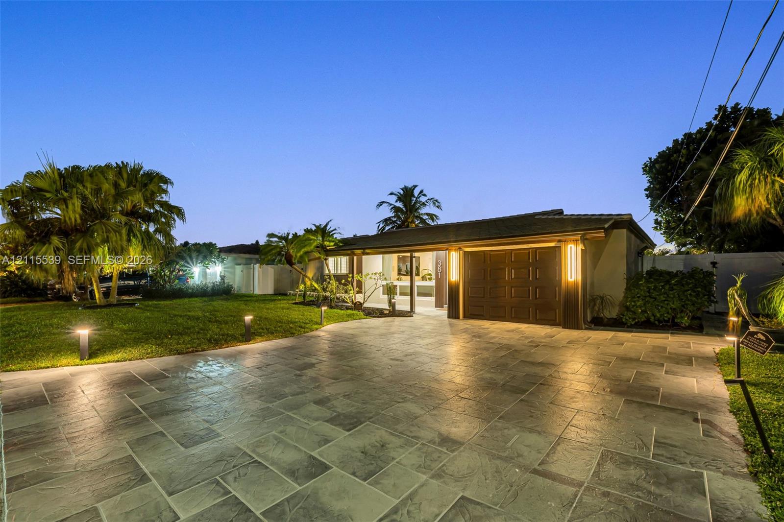 Single-story residence featuring a large stone-paver driveway, integrated garage, manicured lawn, and mature palm trees