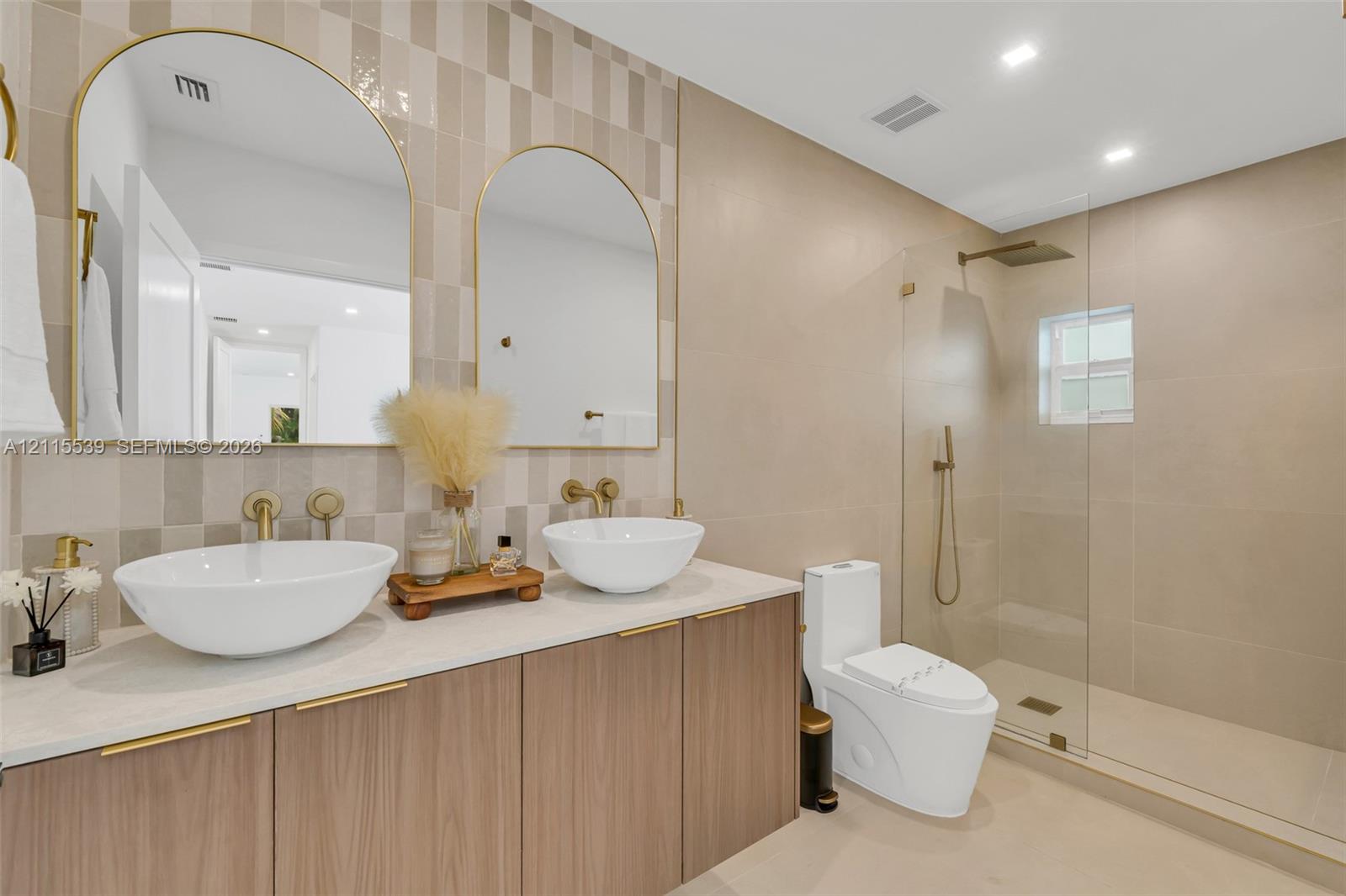 Dual vanity featuring vessel sinks, quartz countertops, wood-finish cabinetry, arched mirrors with gold-toned frames, and a walk-in shower with a glass enclosure