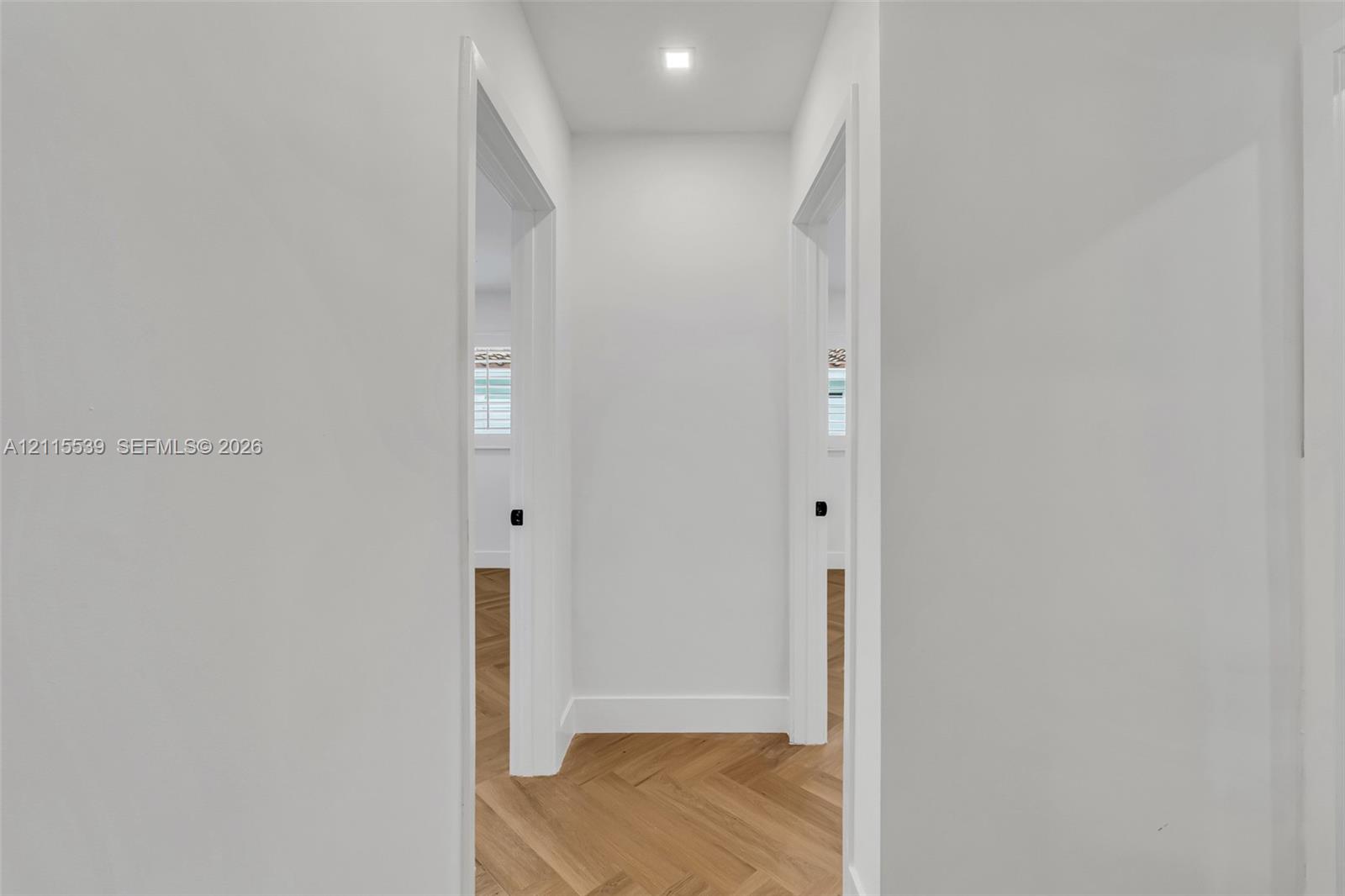 Interior hallway featuring light wood-finish flooring in a herringbone pattern, smooth white walls, recessed ceiling lighting, and matte black door hardware