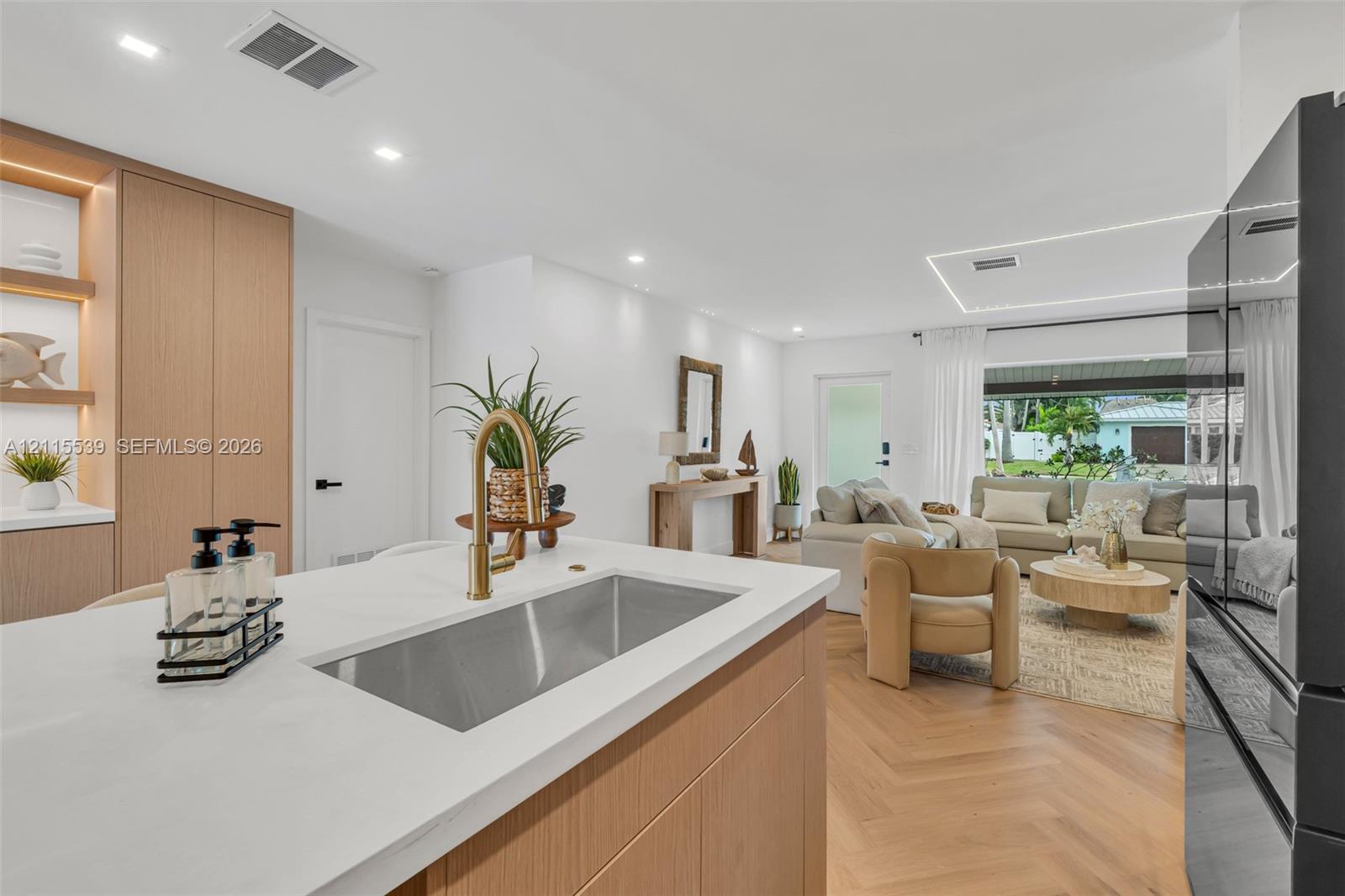 Kitchen island featuring a white countertop and stainless steel undermount sink with a brass-finish faucet, light wood-finish cabinetry, and chevron-pattern wood-finish flooring