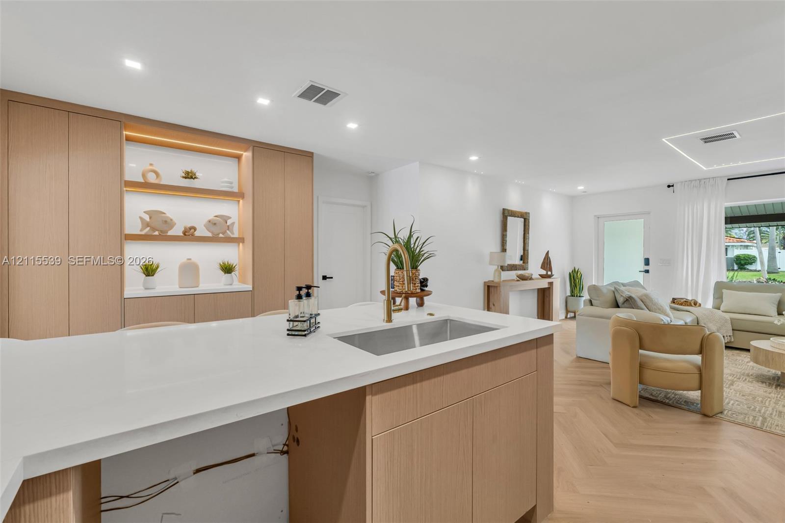 Kitchen featuring light wood-finish cabinetry, a white quartz countertop, an undermount stainless steel sink with a brass faucet, built-in recessed shelving with integrated lighting, and herringbone wood-finish flooring