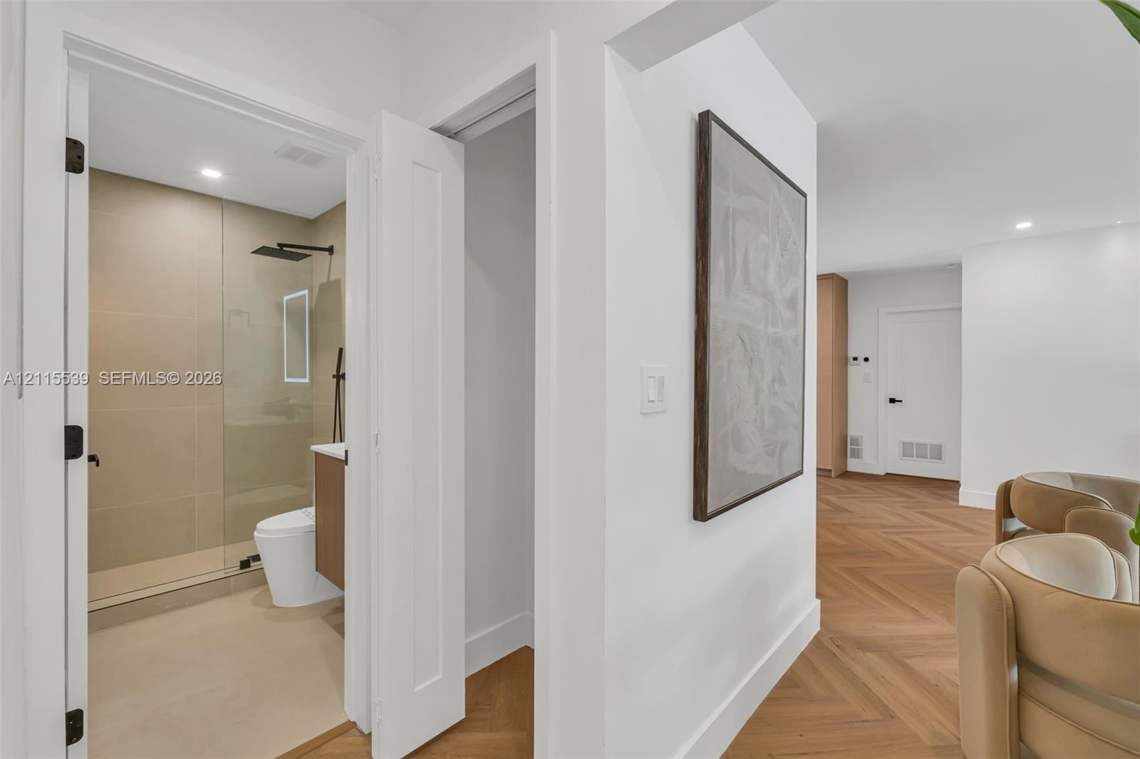 Herringbone wood-finish flooring, walk-in shower with large-format tile, frameless glass shower enclosure, matte black rainfall showerhead, and integrated LED bathroom mirror