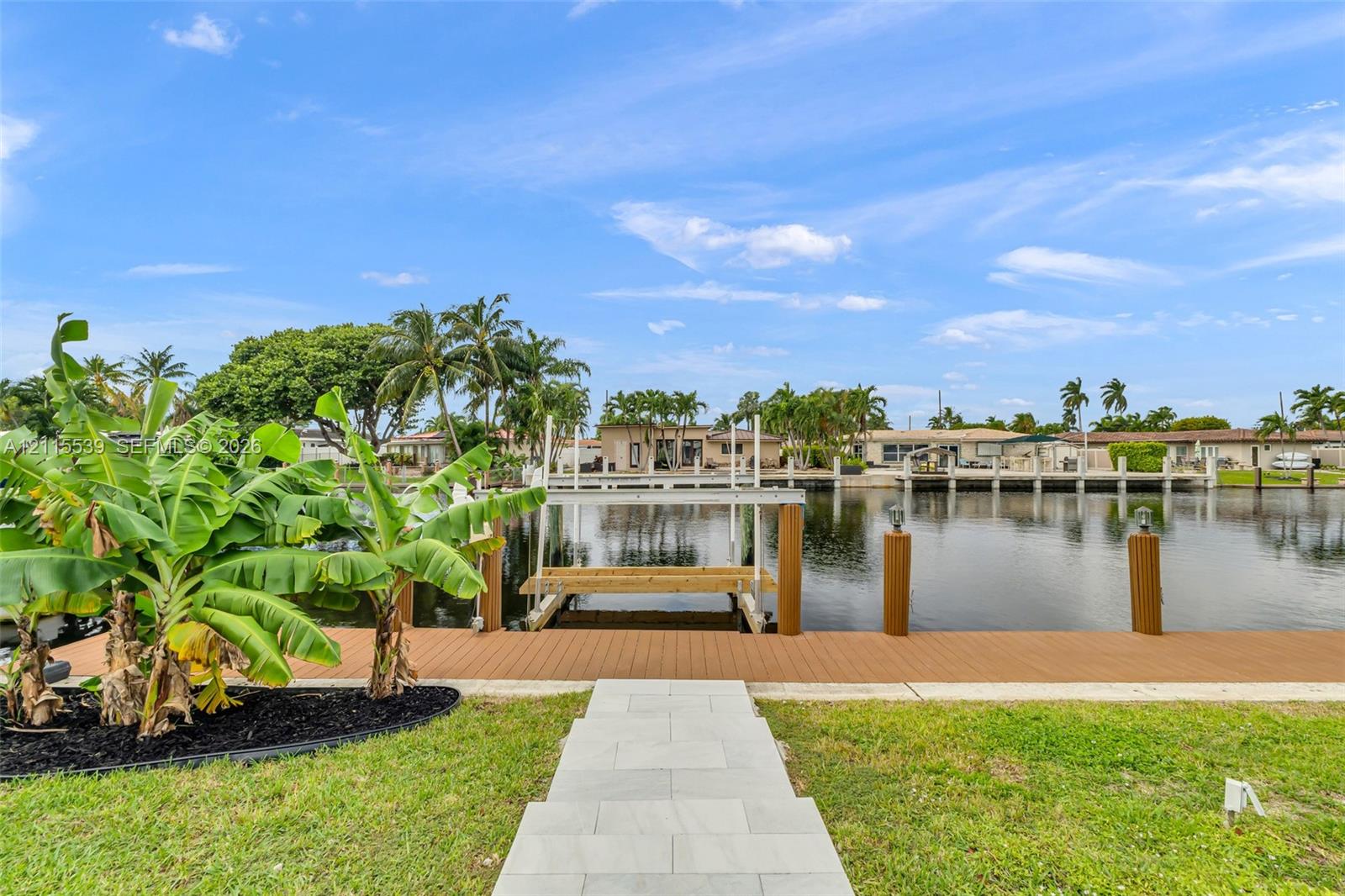 Waterfront property featuring a composite deck, private boat lift, and anchored wooden pilings, complemented by a paver stone walkway and tropical landscaping