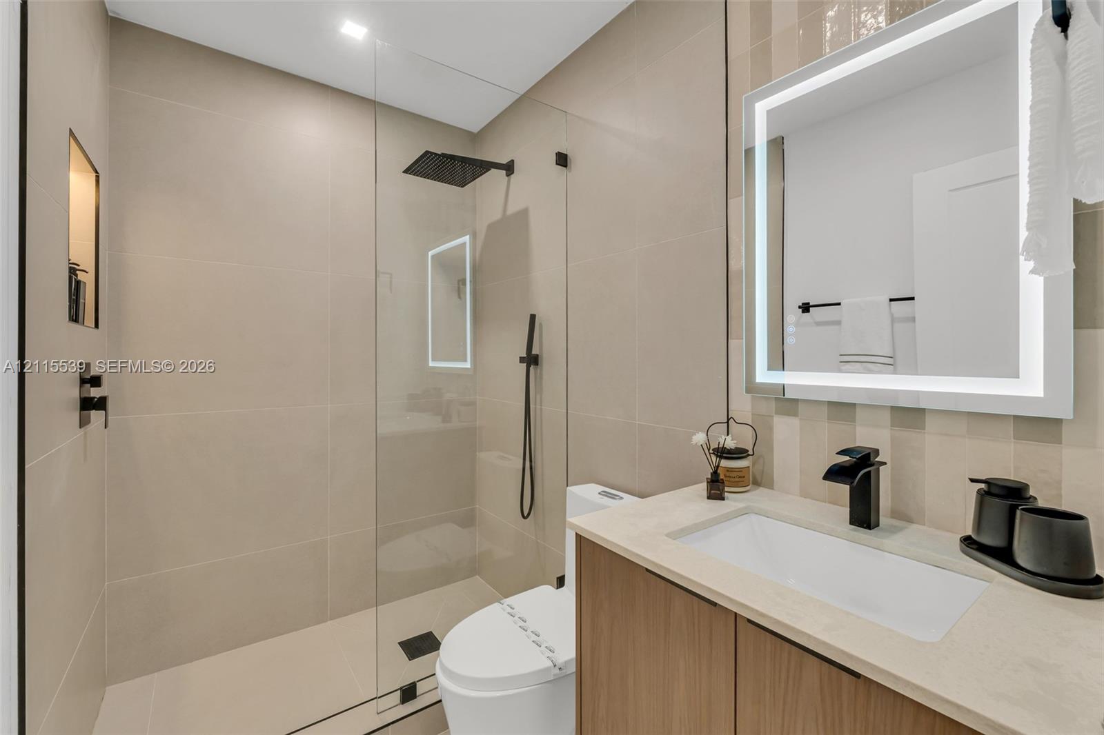 Bathroom featuring a wood-finish vanity with a light-toned stone countertop, a glass-enclosed walk-in shower with matte black fixtures, a built-in shower niche, and an integrated LED-backlit mirror