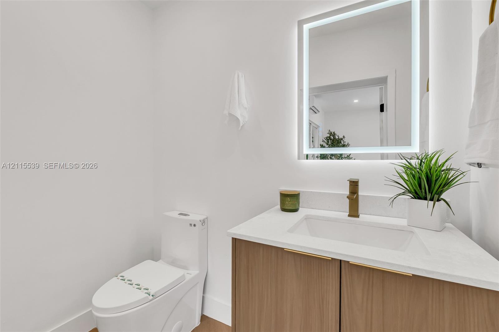 Wood-finish vanity cabinet, white solid-surface countertop, rectangular undermount sink, brass-tone faucet, and rectangular LED-backlit mirror