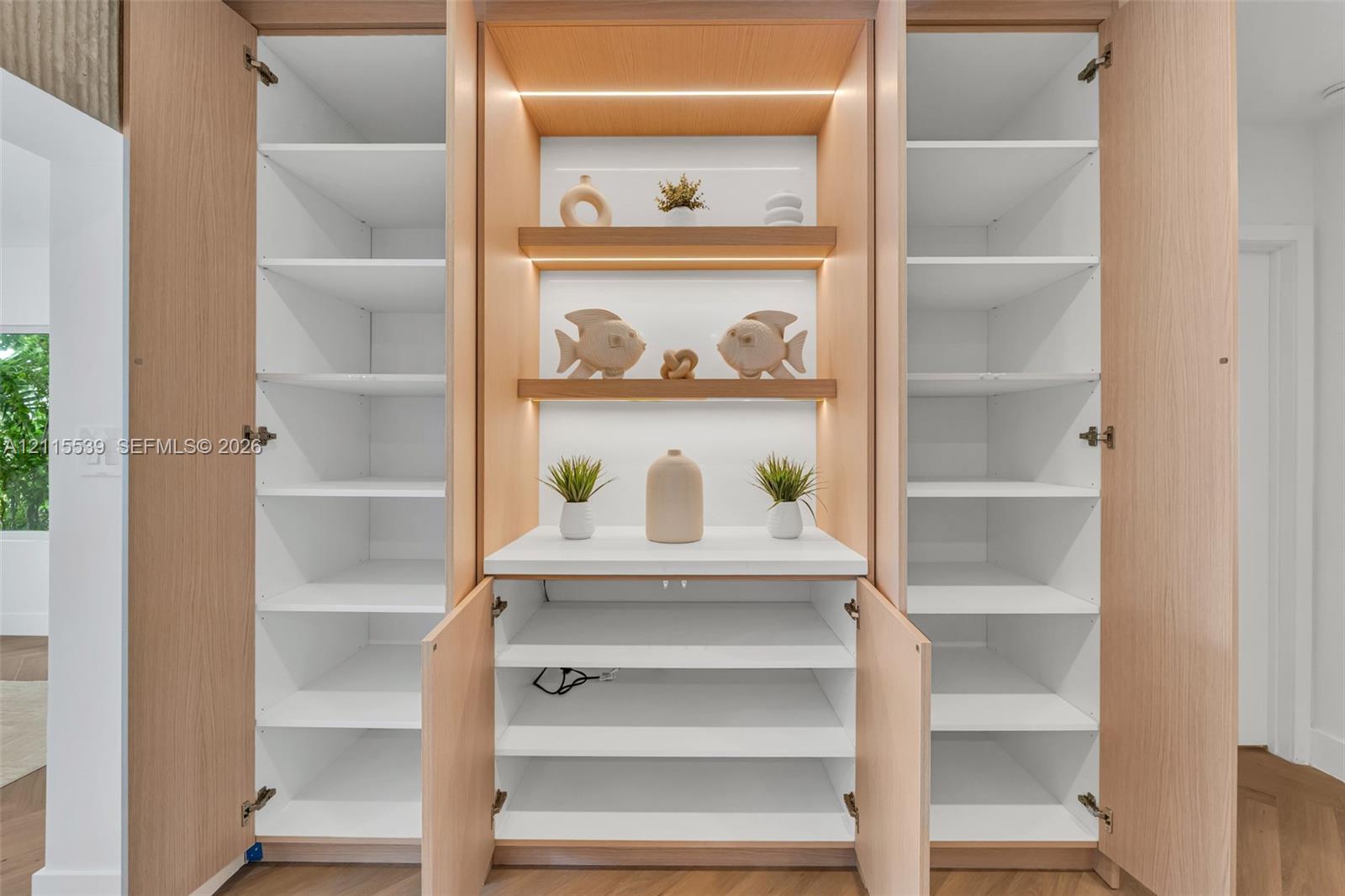 Custom built-in cabinetry featuring light wood-finish veneer, integrated LED shelf lighting, fixed white shelving, and a central solid-surface countertop
