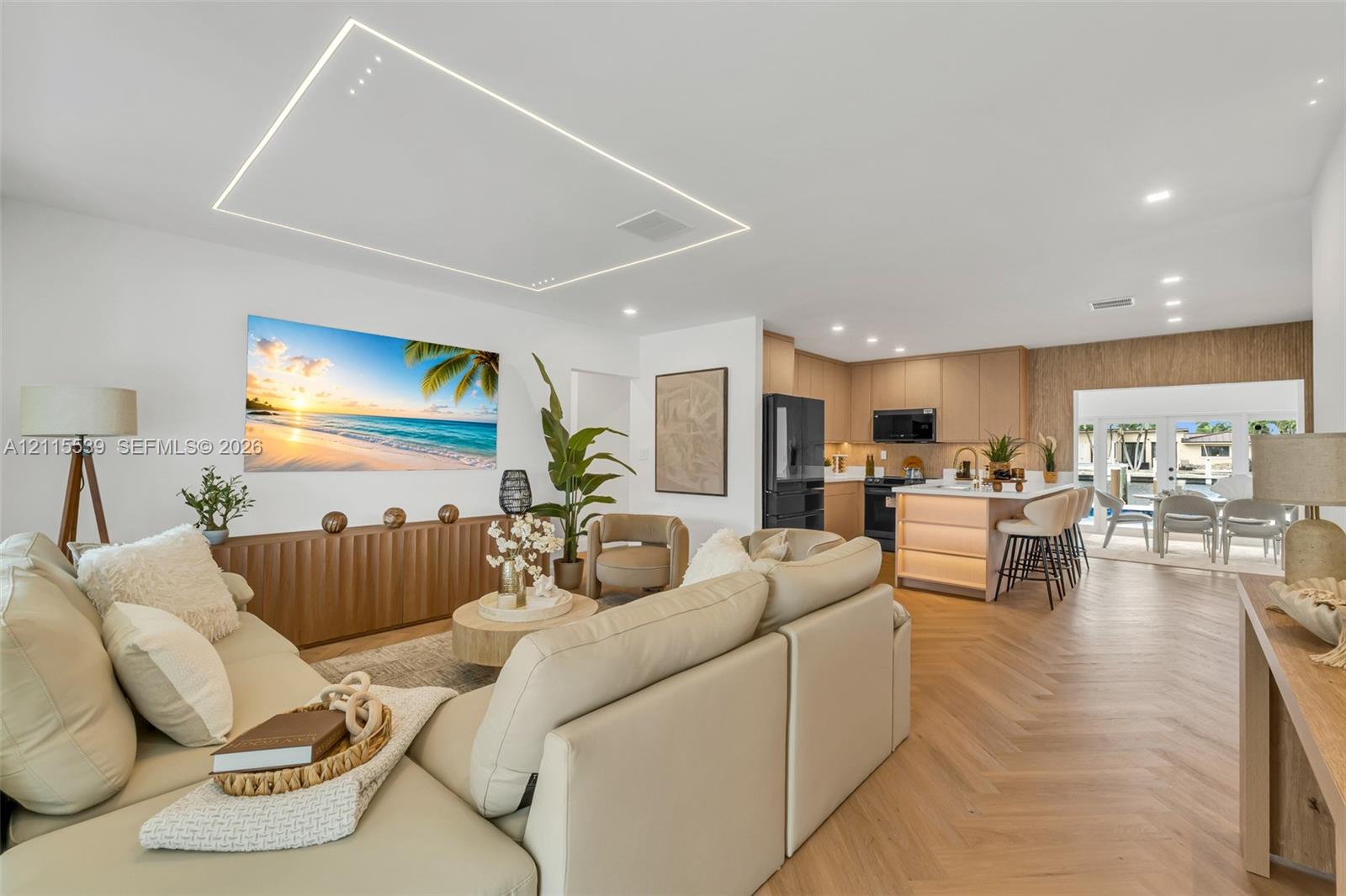 Open-concept living space featuring wood-finish flooring in a herringbone pattern, a recessed rectangular tray ceiling with perimeter lighting, recessed ceiling spotlights, a kitchen island with integrated under-counter lighting, and French doors