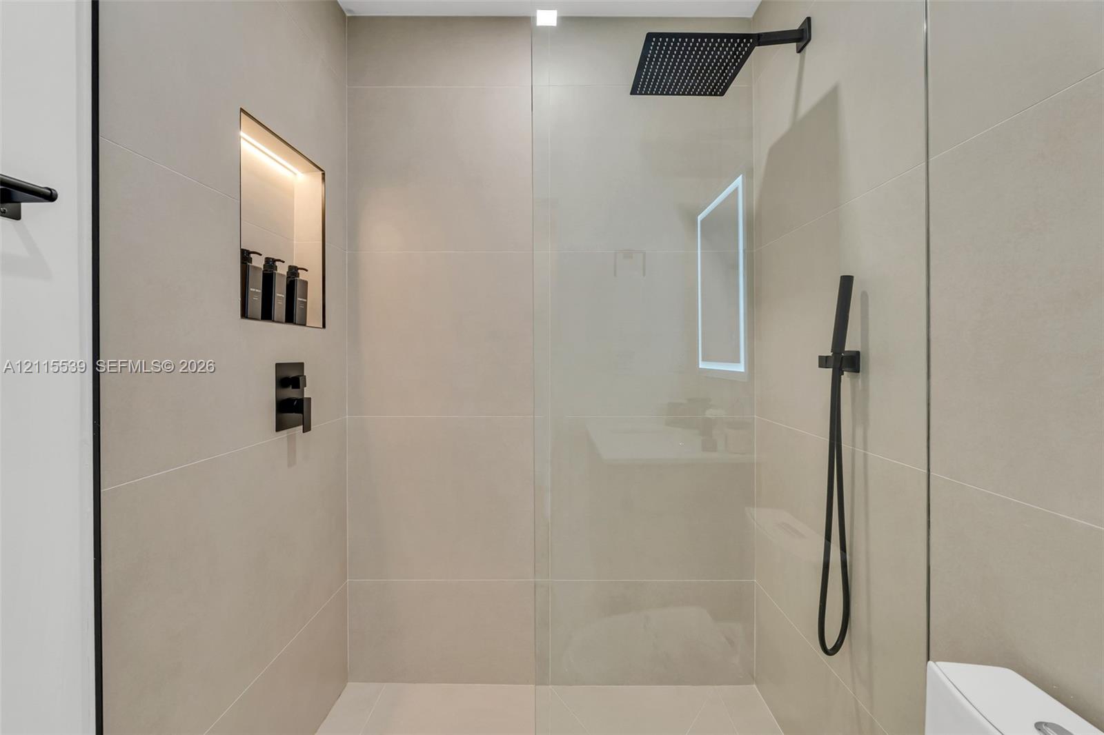 Walk-in shower featuring large-format neutral tile, frameless glass enclosure, matte black rainfall showerhead, integrated handheld sprayer, and recessed tiled niche with lighting