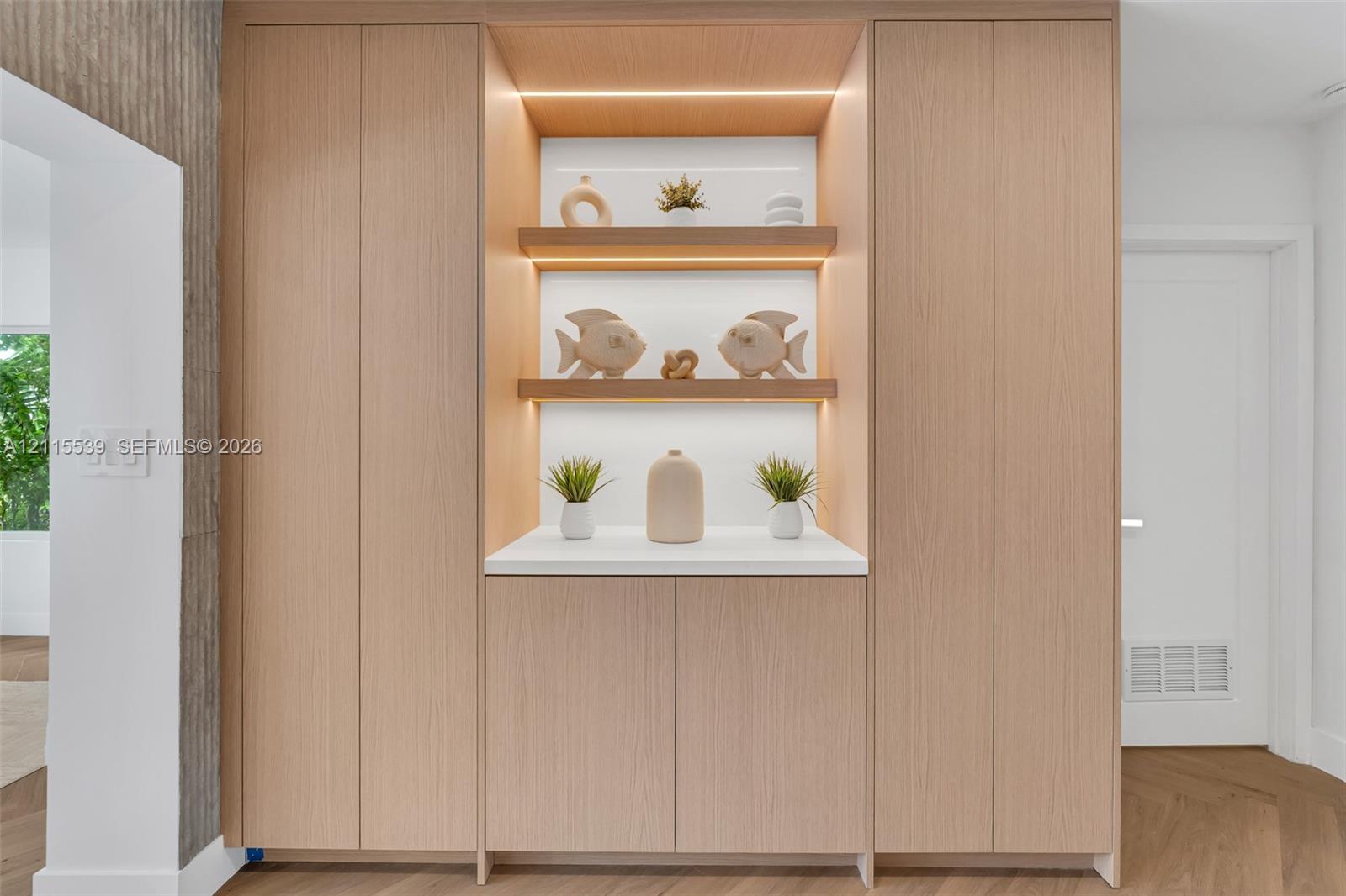 Floor-to-ceiling cabinetry with light wood-finish veneer, integrated recessed display shelving, built-in task lighting, white solid-surface countertop, and herringbone wood-finish flooring