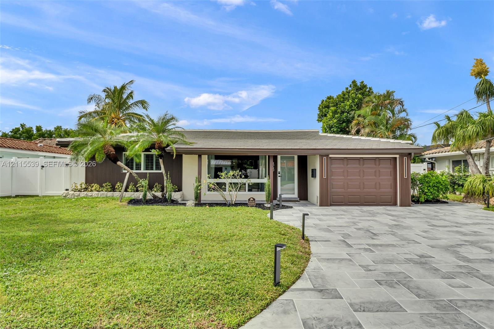 Single-story exterior featuring a stone-paver driveway, attached garage, large picture window, and landscaped lawn with palm trees