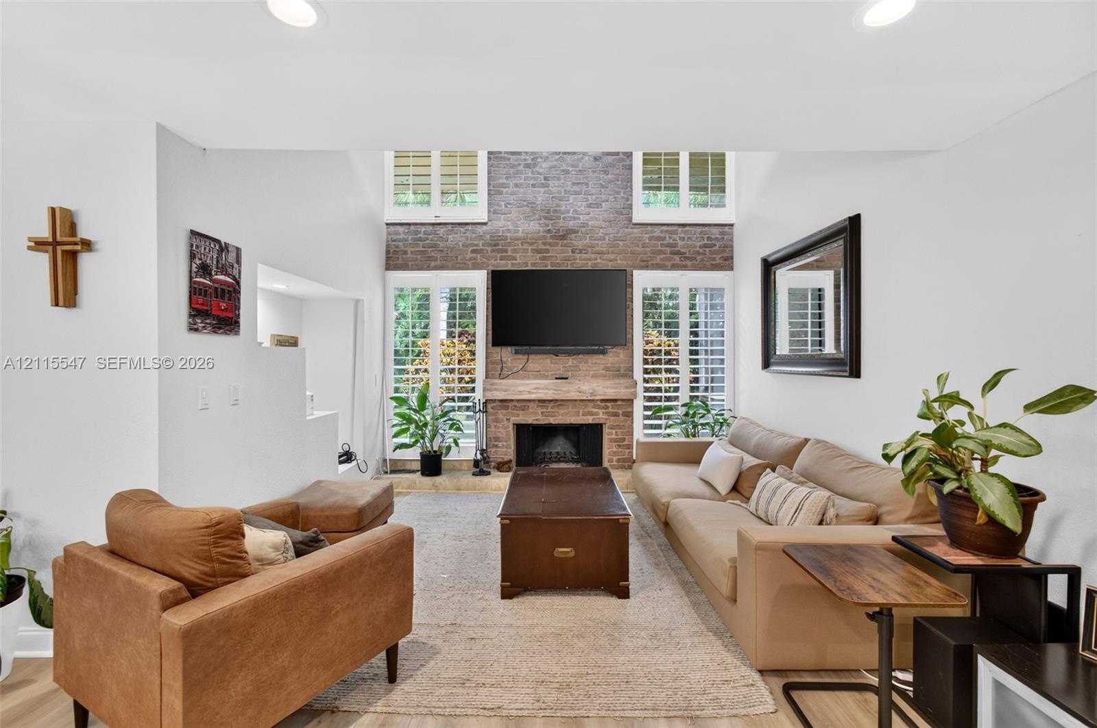 Double-height living room featuring a floor-to-ceiling brick fireplace, recessed lighting, and plantation shutters flanking multi-pane glass doors