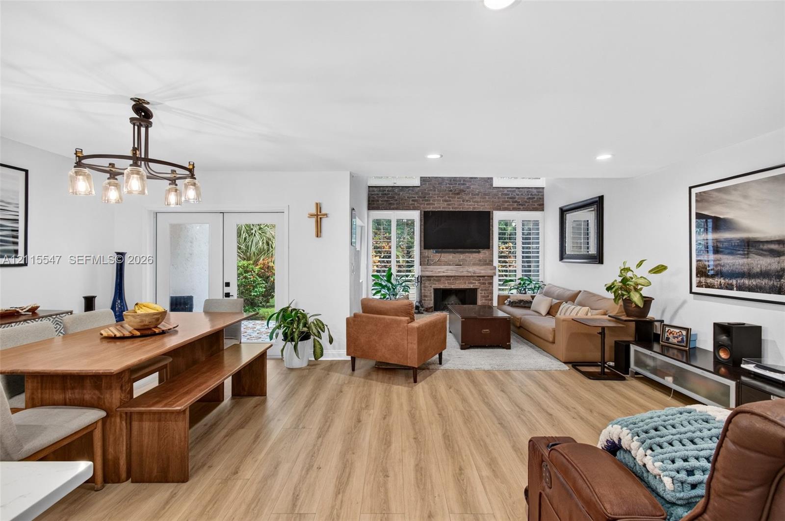 Open-concept interior featuring wood-finish flooring, a full-height brick fireplace, recessed lighting, a multi-arm chandelier, and plantation shutters