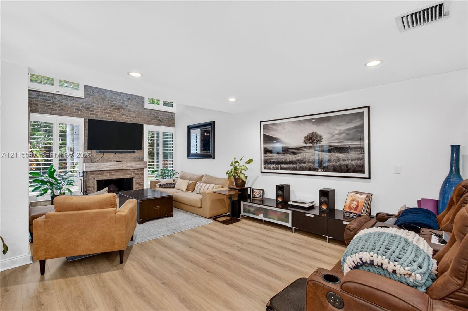 Living area featuring wood-finish flooring, floor-to-ceiling brick fireplace surround, natural wood mantel, and white plantation shutters