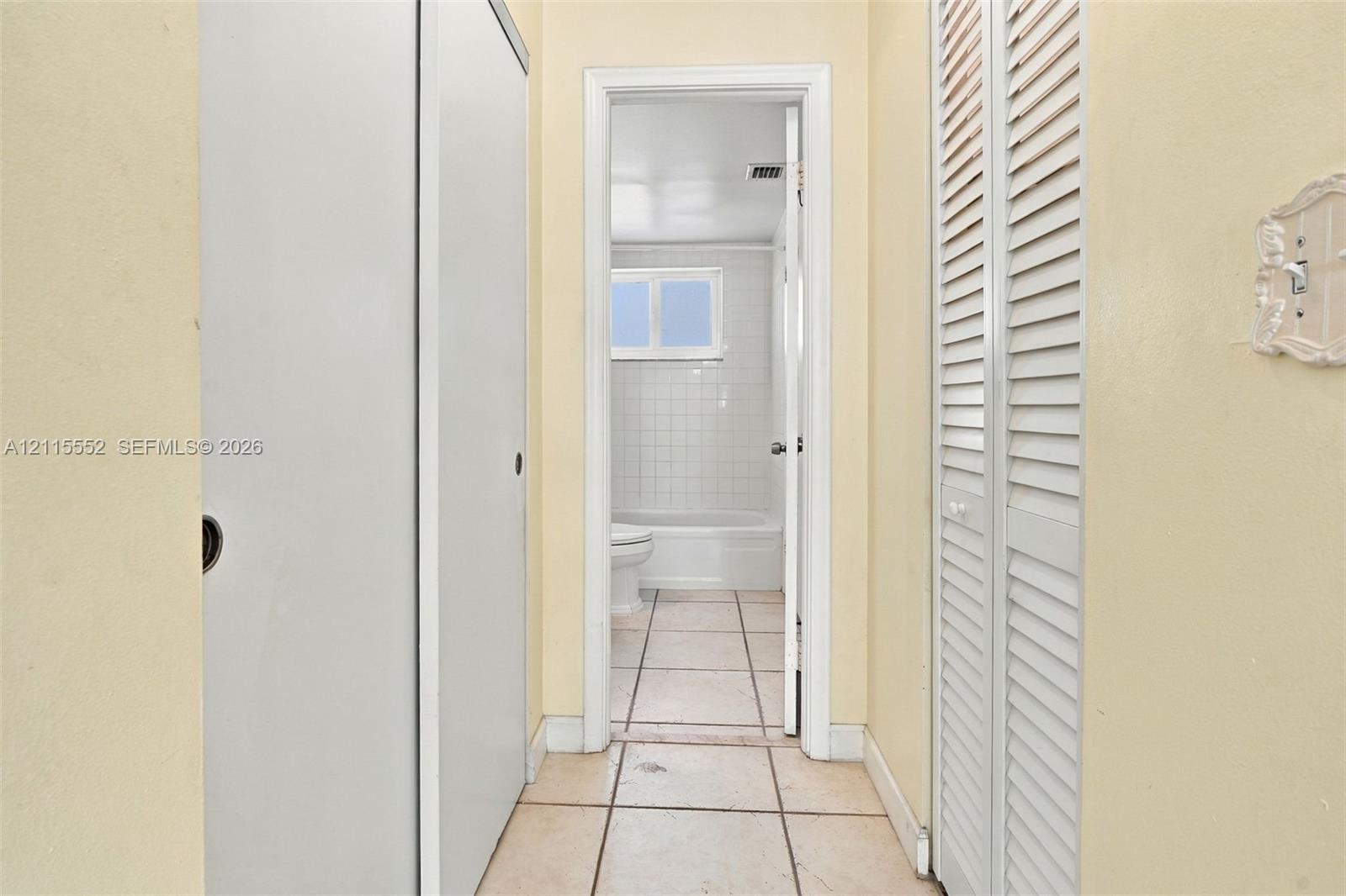 Hallway featuring dual sets of white louvered closet doors, tile flooring, and a doorway revealing a tiled tub surround and window