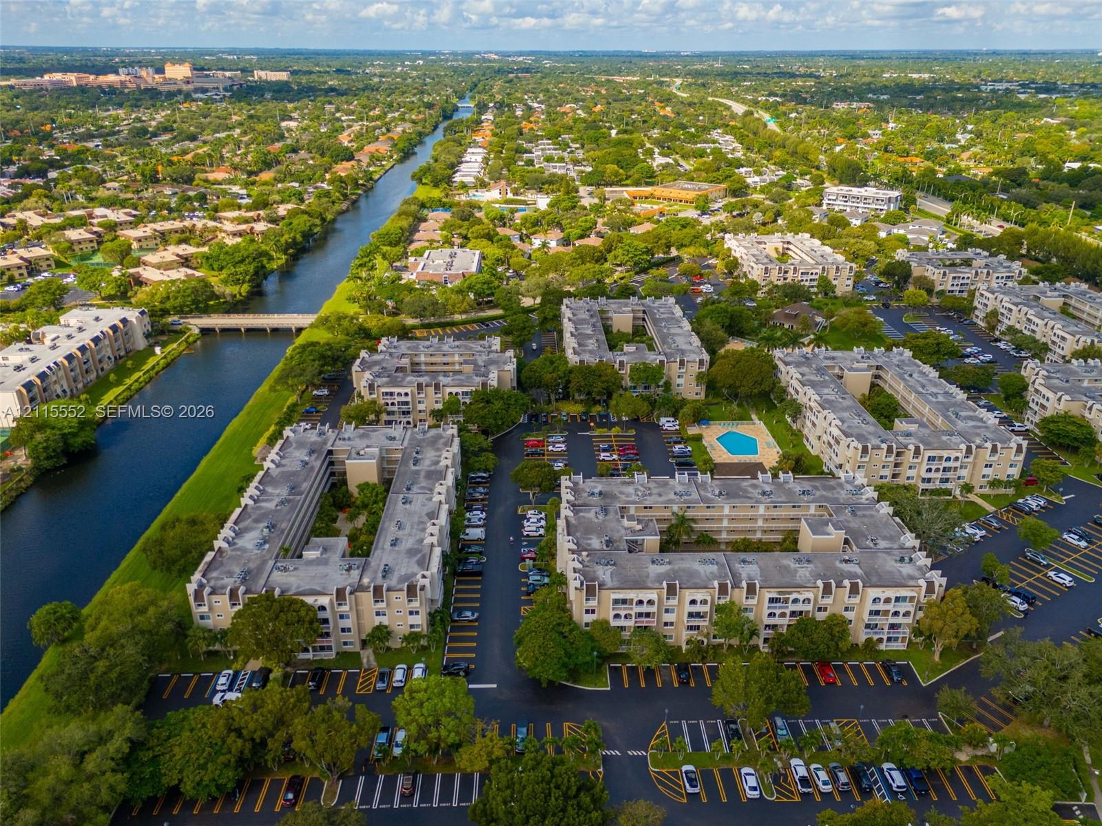 Aerial perspective of a multi-building residential complex featuring a swimming pool, surface parking, and canal frontage with mature landscaping