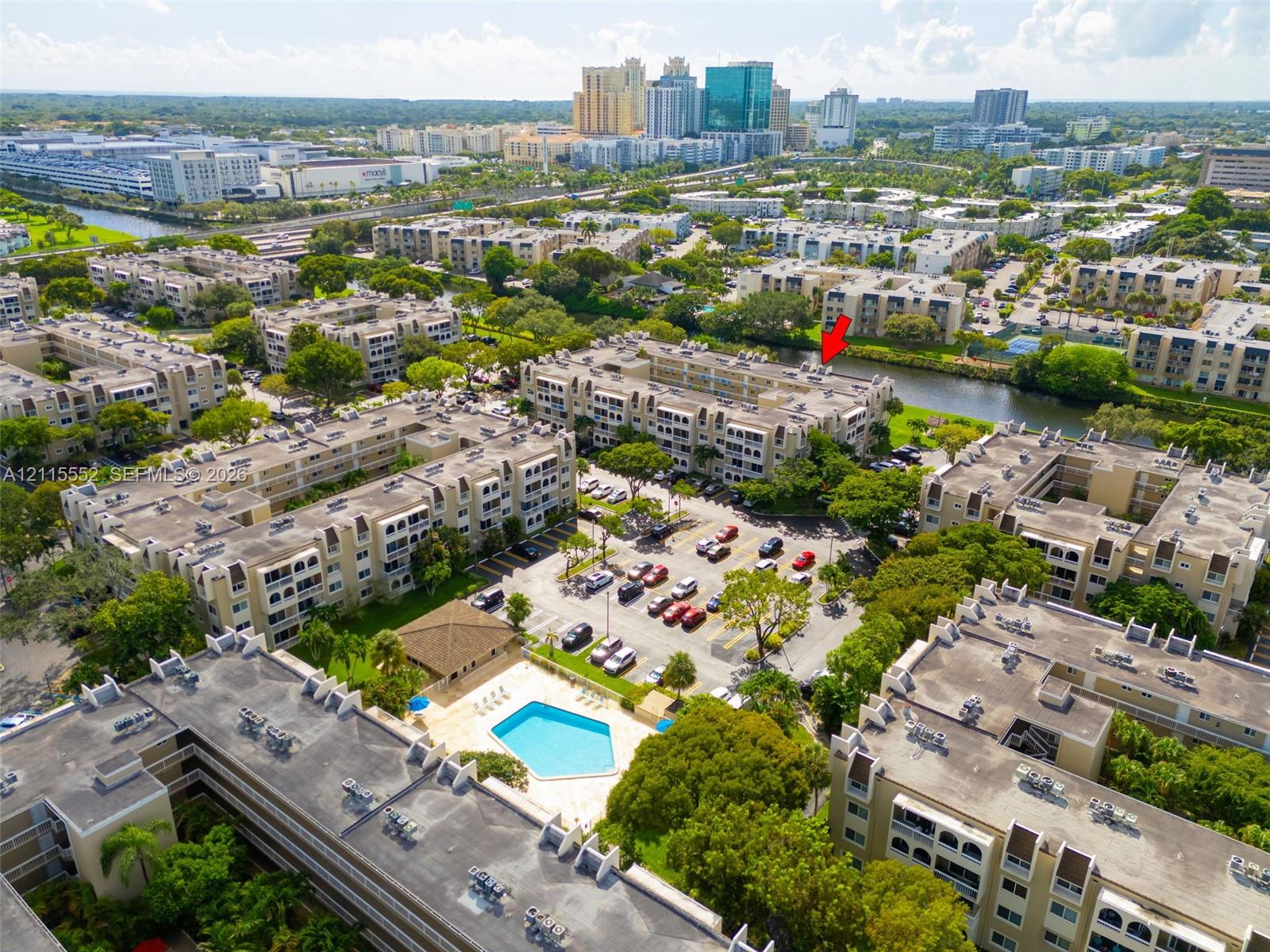 Aerial perspective of a multi-story residential complex featuring an outdoor swimming pool, paved surface parking, and waterfront grounds