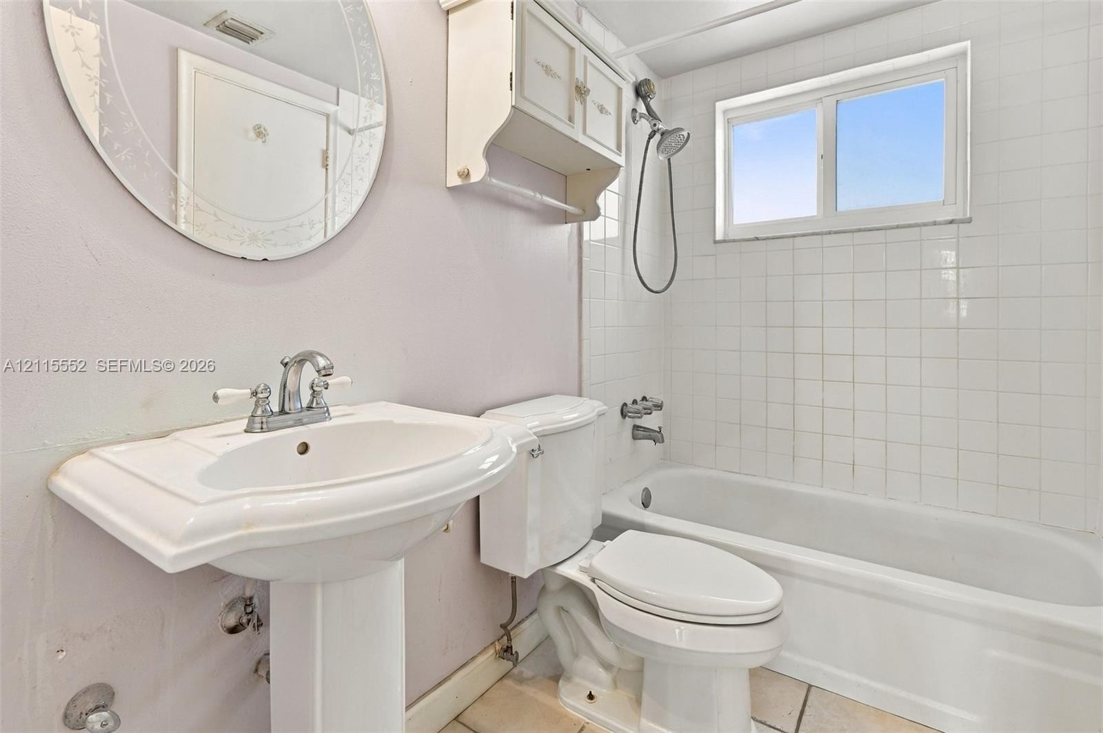 Pedestal sink with chrome dual-handle faucet, oval mirror, wall-mounted storage cabinet, and white tile-surround bathtub featuring a handheld showerhead
