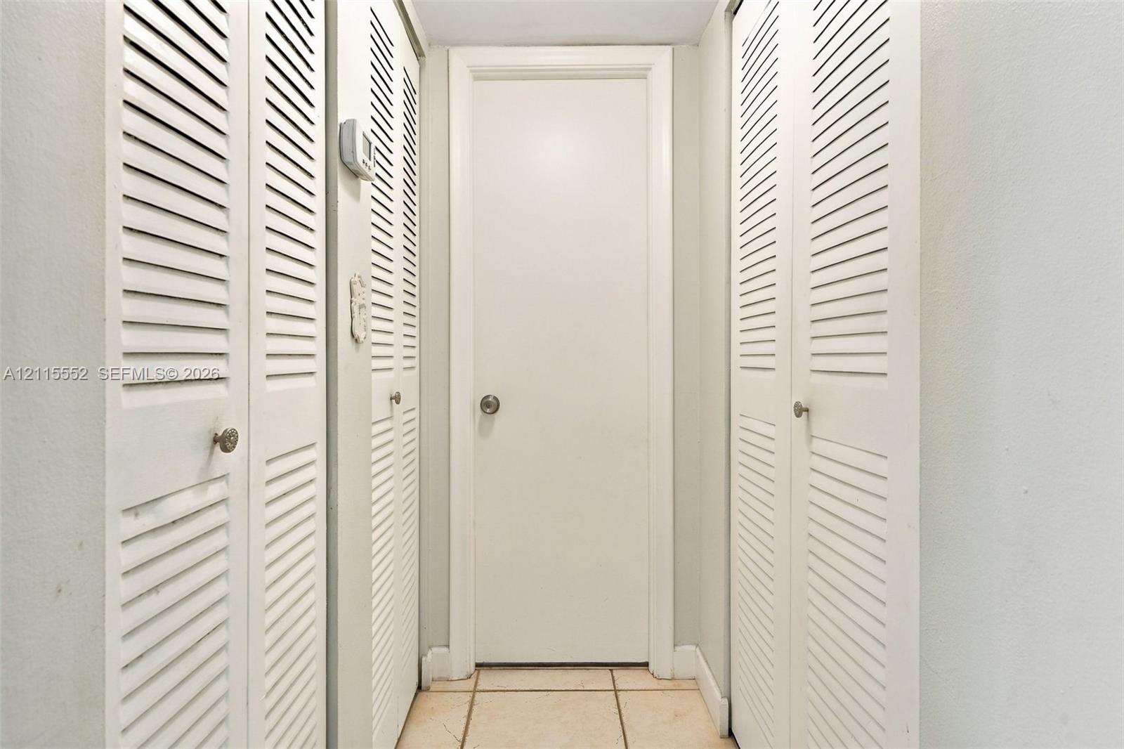 Hallway featuring dual sets of white louvered doors, a solid-panel door with a metallic knob, and light-toned tile flooring