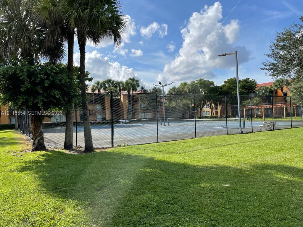 Fenced tennis court with overhead lighting, manicured green space, and mature palm trees