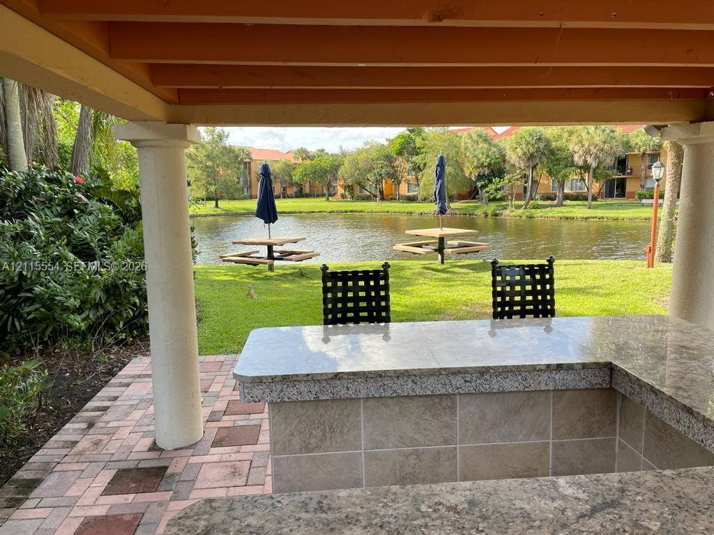 Covered outdoor kitchen featuring a stone-tile facade, stone countertop, and brick paver flooring, situated under a timber-framed pergola with structural columns overlooking a pond