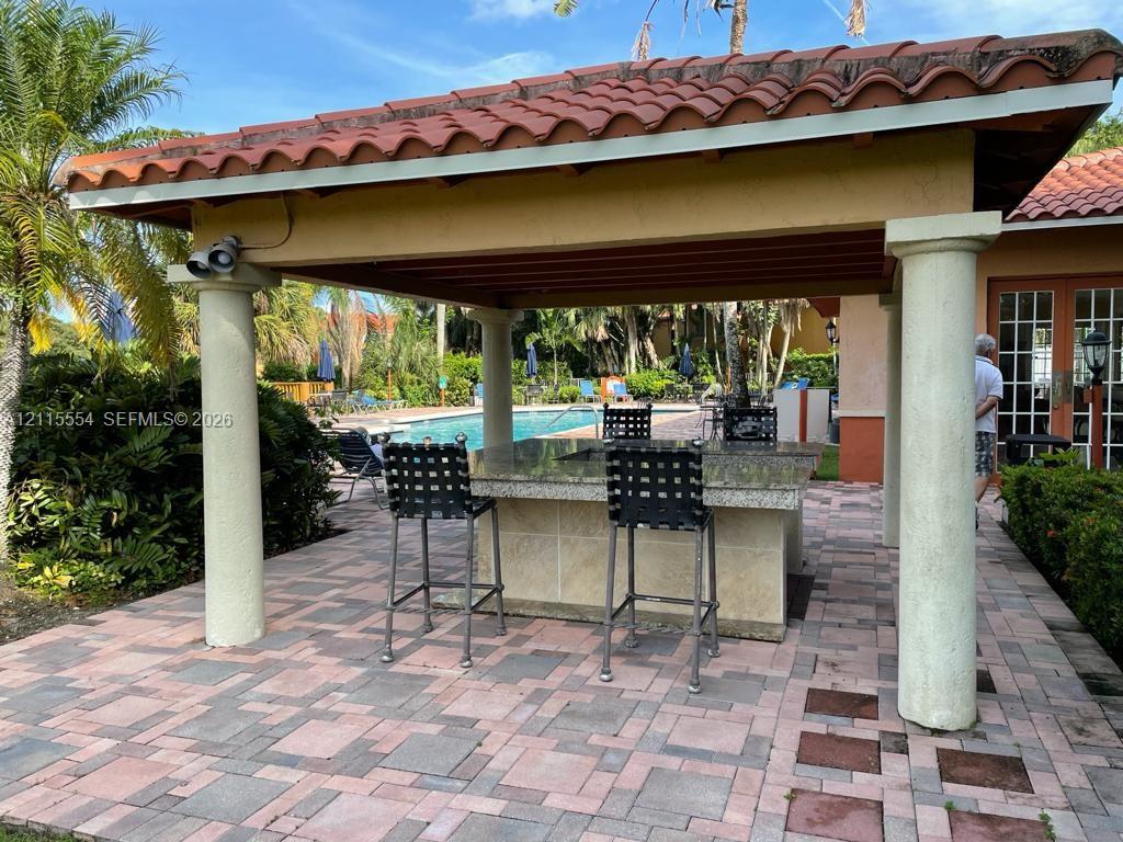 Covered outdoor kitchen featuring a stone-topped island, paver patio flooring, and classical columns under a terracotta-tiled roof