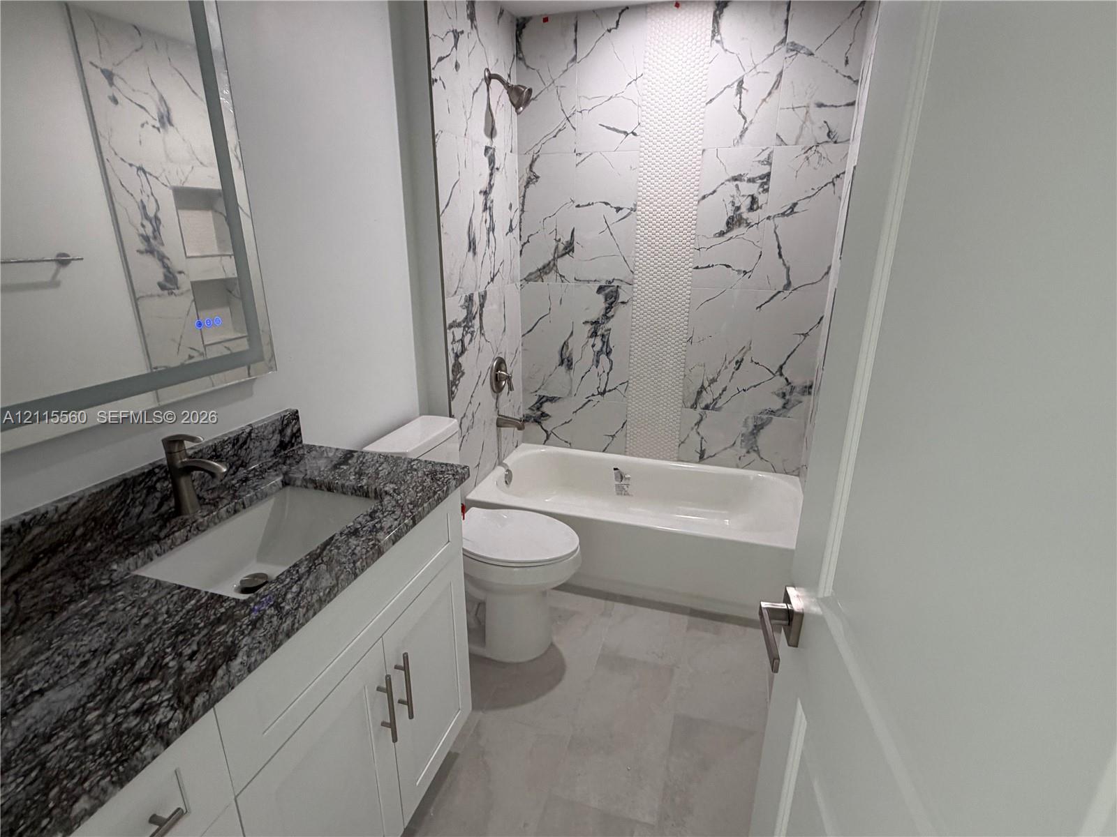 Bathroom featuring a vanity with stone-finish countertop, under-mount sink, white cabinetry with brushed metal hardware, and a bathtub surround with patterned wall tile and an integrated recessed niche
