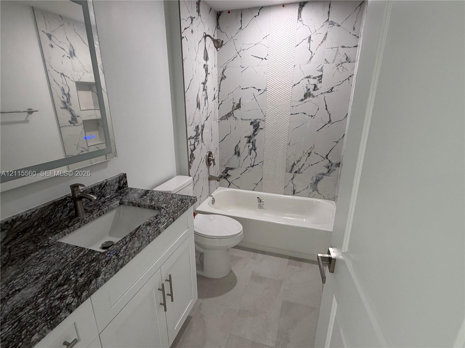 Bathroom featuring a vanity with stone-finish countertop, undermount sink, porcelain tile tub surround with an integrated shower niche, and rectangular floor tiling