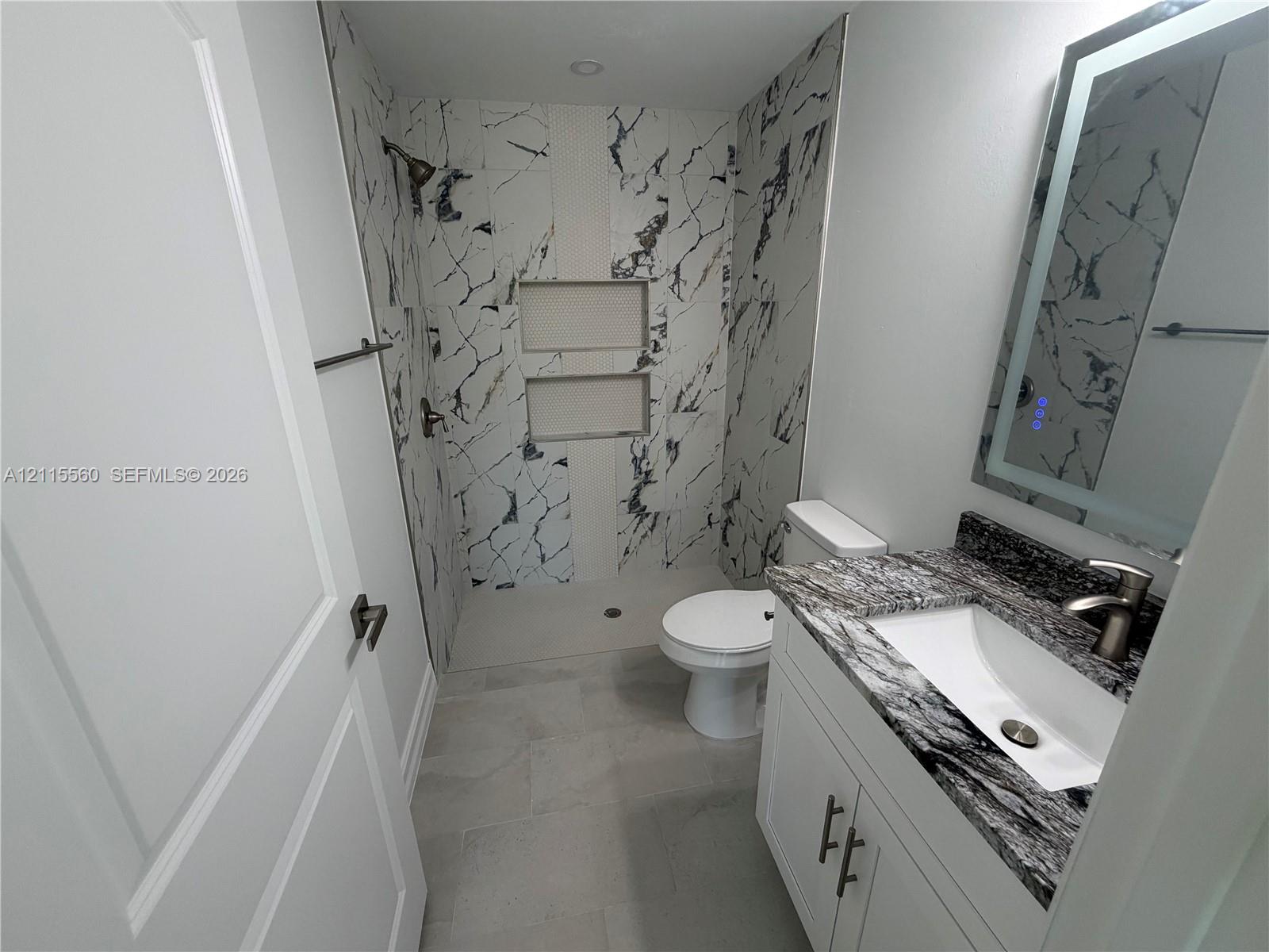Bathroom featuring a shower with dual built-in niches, stone-patterned tile surround, vanity with a decorative stone countertop, undermount sink, and integrated LED-lit mirror