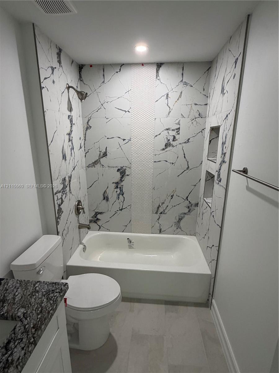 Bathroom featuring white and grey large-format stone-look wall tiles with a central mosaic vertical accent, built-in dual recessed shower shelving, a white acrylic tub, and a vanity with a dark-patterned stone-look countertop
