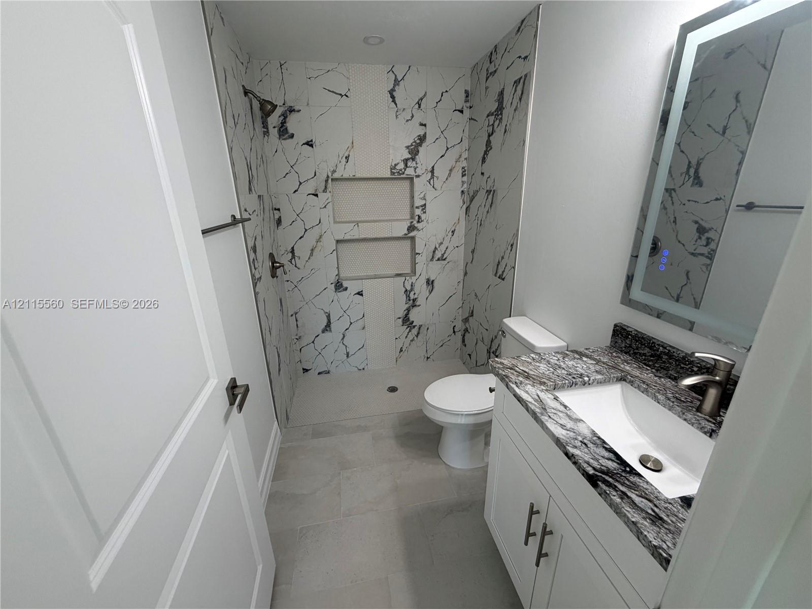 Bathroom featuring a stone-surface vanity with an integrated sink, illuminated vanity mirror, walk-in shower with dual recessed niches, and large-format tile flooring
