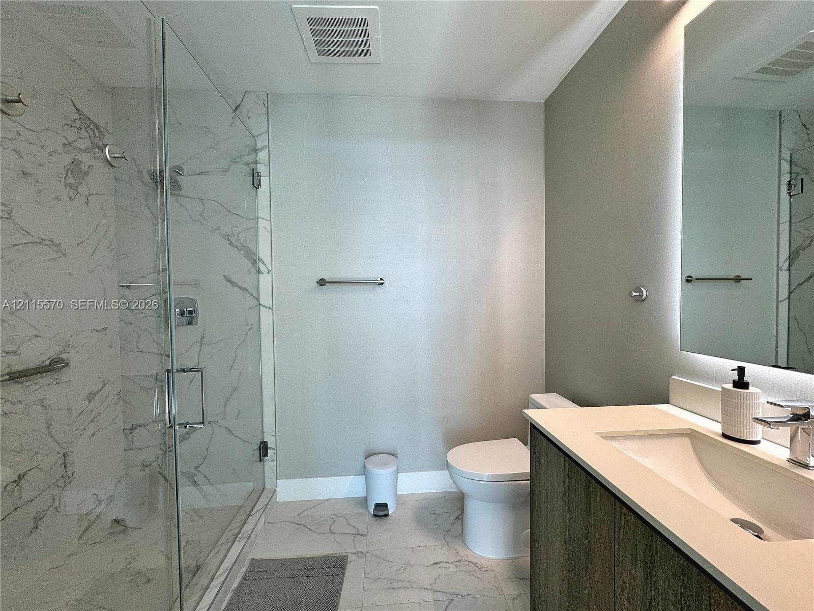 Walk-in shower with floor-to-ceiling stone-look tile, frameless glass enclosure, wood-finish vanity with a quartz countertop, and a backlit wall mirror