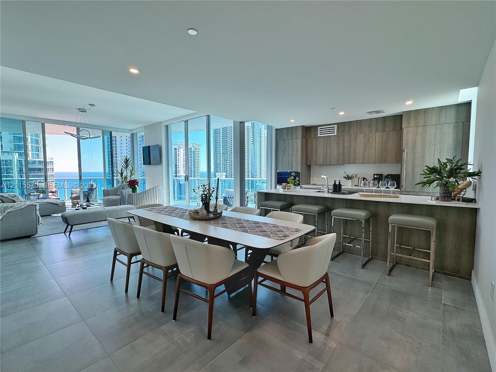 Open-concept living space featuring expansive floor-to-ceiling glass doors, wood-finish cabinetry, a large kitchen island with integrated seating, and neutral stone-finish flooring