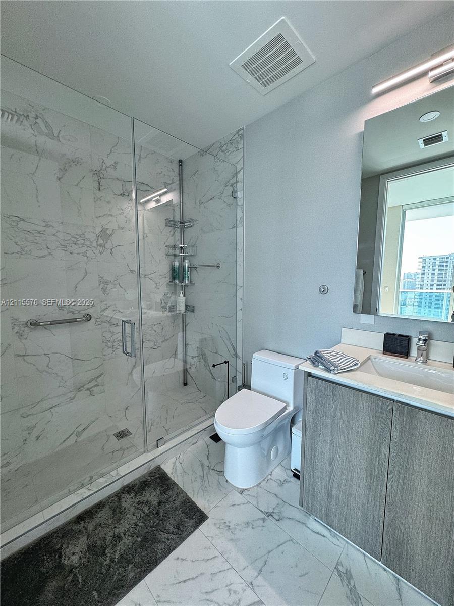 Bathroom featuring a glass-enclosed walk-in shower with floor-to-ceiling stone-finish tile, vanity with integrated basin and wood-finish cabinetry, and oversized wall mirror