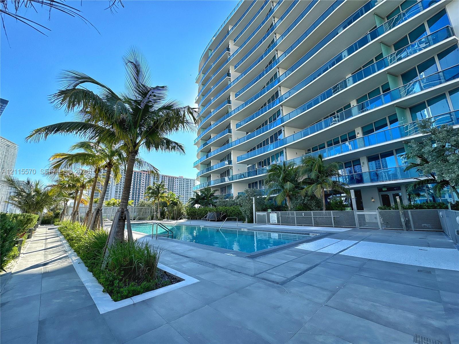 Multi-story glass-facade residential tower featuring private balconies, a rectangular swimming pool, large-format tile decking, and palm-lined landscaping
