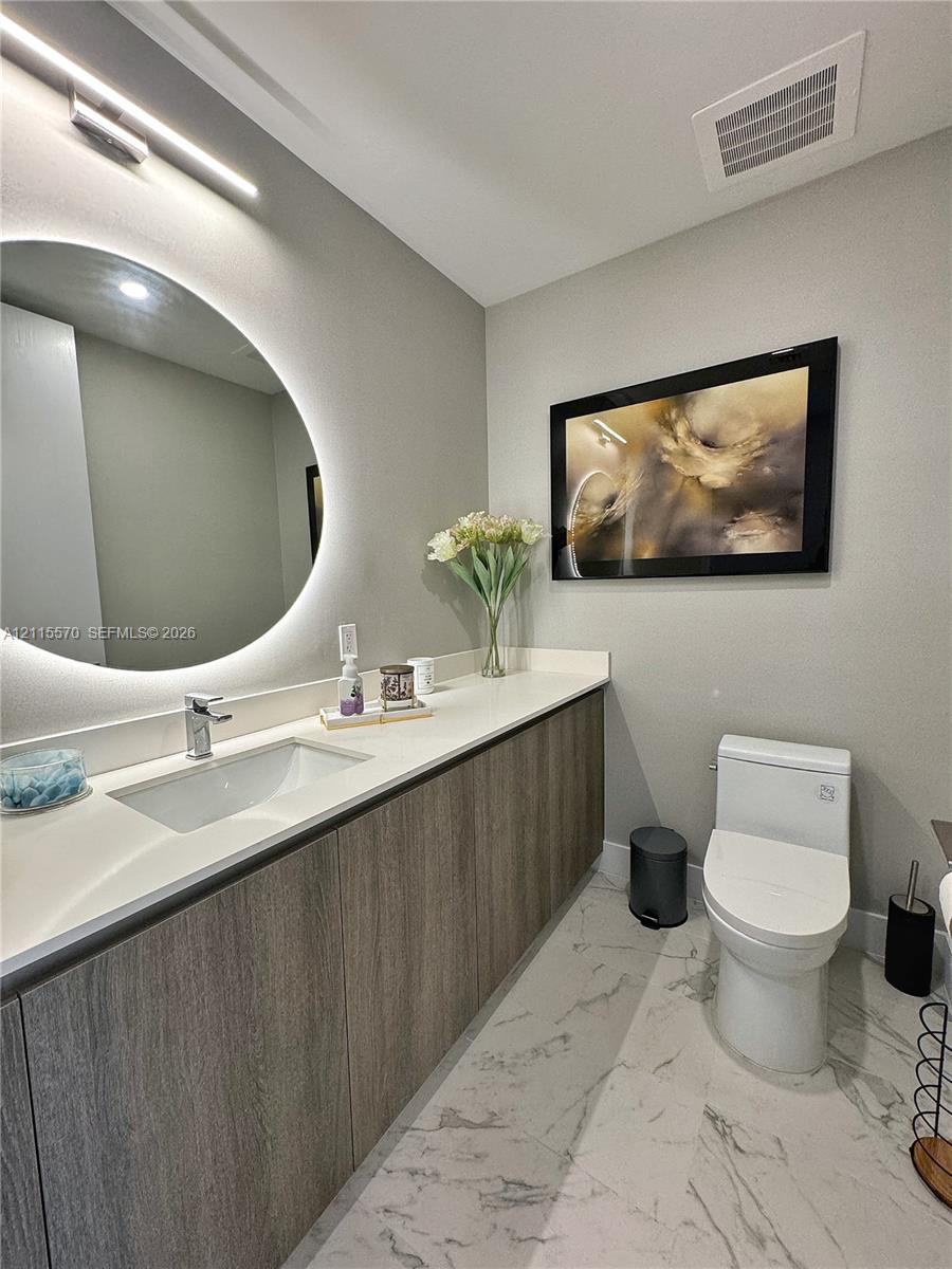Modern bathroom featuring a wood-finish floating vanity, integrated stone countertop with an undermount sink, LED-backlit circular mirror, and large-format veined tile flooring
