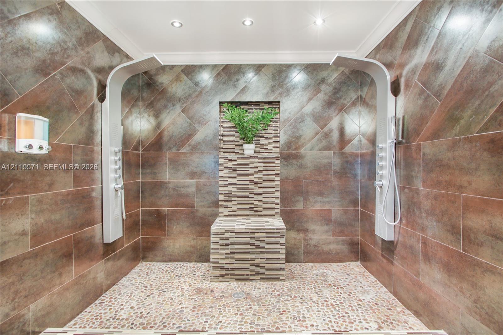 Walk-in shower featuring large-format metallic-finish wall tiles, dual vertical shower panels with integrated body sprayers, a central stone-tiled bench, and natural pebble flooring