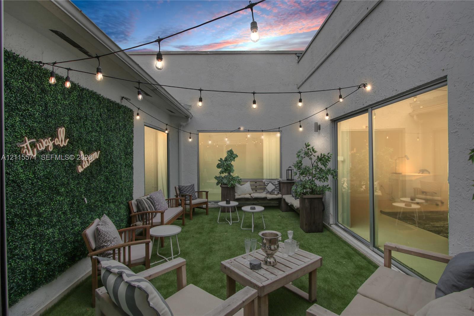 Private courtyard featuring stucco walls, synthetic turf, integrated artificial greenery panel, and overhead string lighting, enclosed by floor-to-ceiling glass paneling