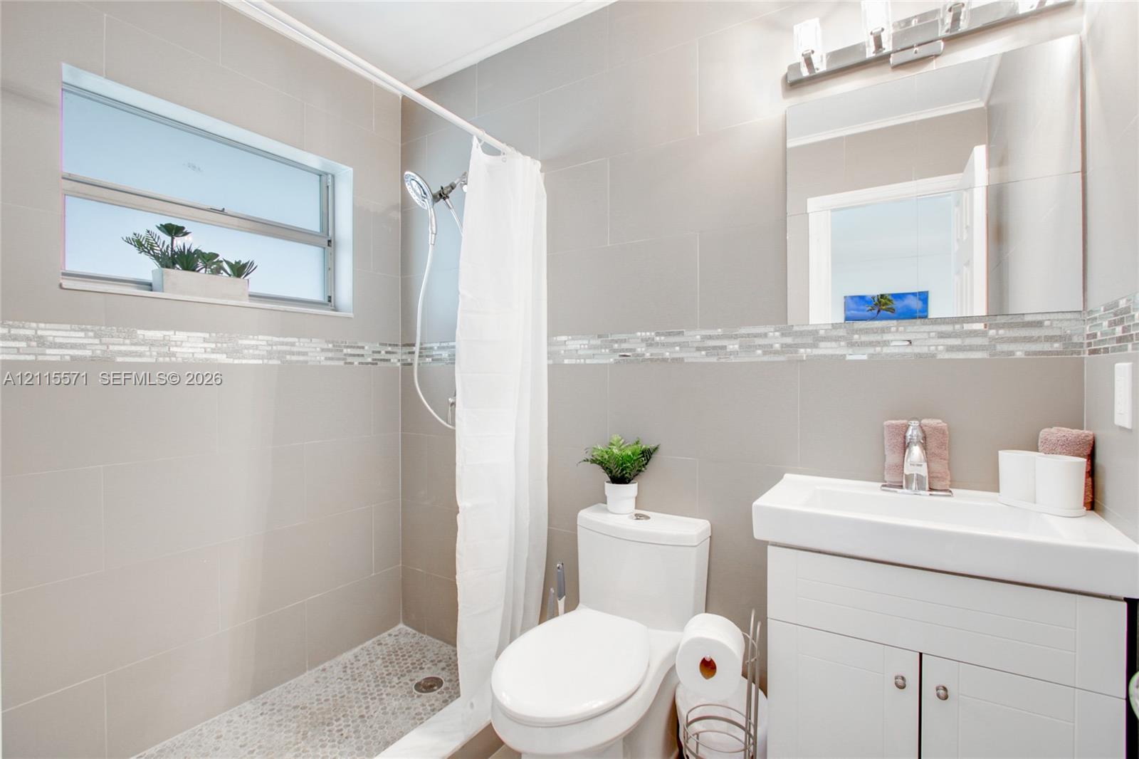 Bathroom featuring large-format wall tile, glass mosaic accent border, pebble-style shower floor tiling, vanity with an integrated sink, and an expansive wall-mounted mirror
