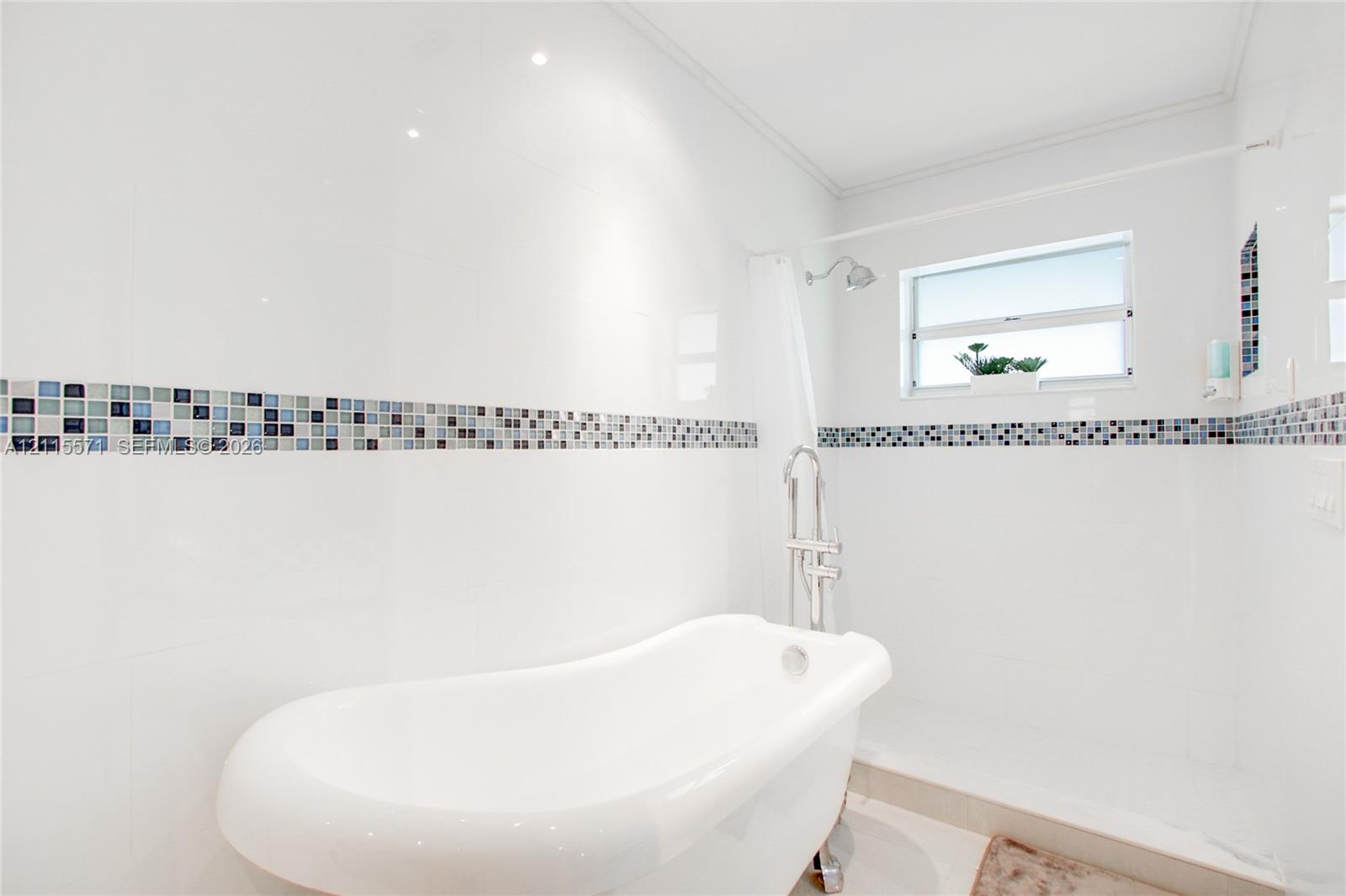 Freestanding soaking tub with a chrome floor-mounted faucet, crisp white wall tile, decorative multi-tonal glass tile inlay, and recessed ceiling lighting