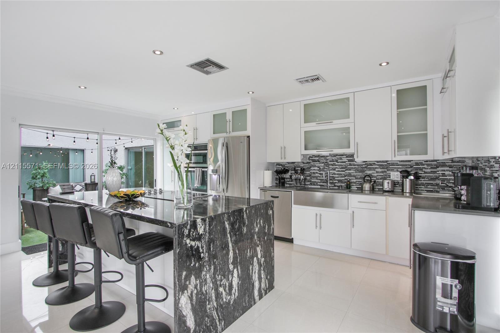 Kitchen featuring white cabinetry, stone-topped central island with waterfall edge, stainless steel farmhouse sink, tiled backsplash, and recessed lighting