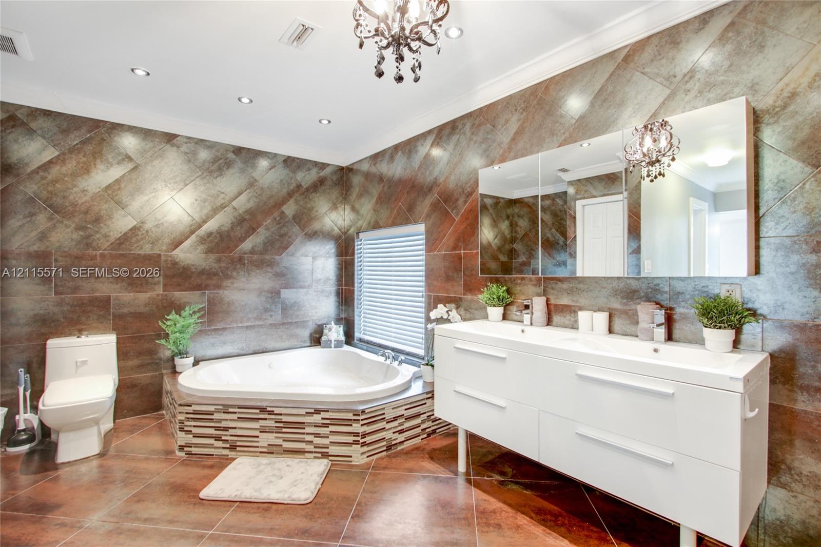 Corner soaking tub with tiled surround, dual-sink white floating vanity, expansive wall-to-wall mirror, metallic-finish wall tiling, and decorative chandelier lighting