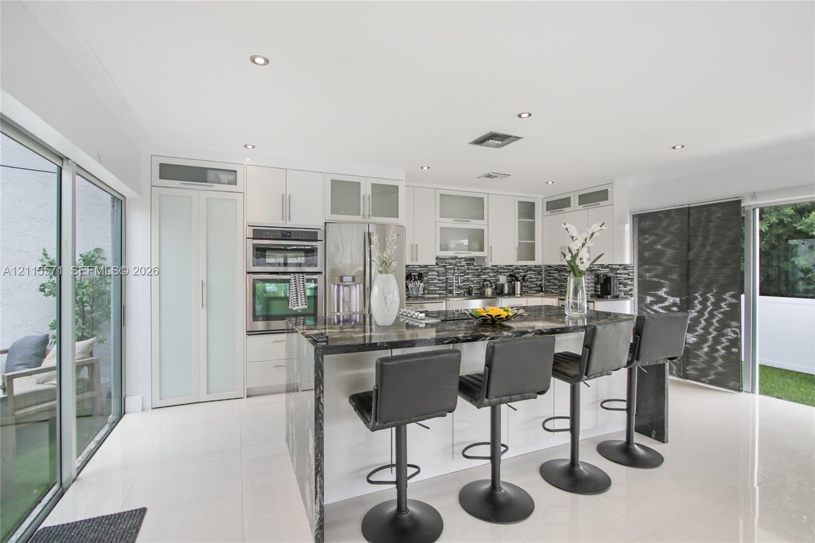 Kitchen featuring white cabinetry with glass-panel accents, stone-topped central island, built-in stainless steel double ovens, patterned tile backsplash, and polished tile flooring