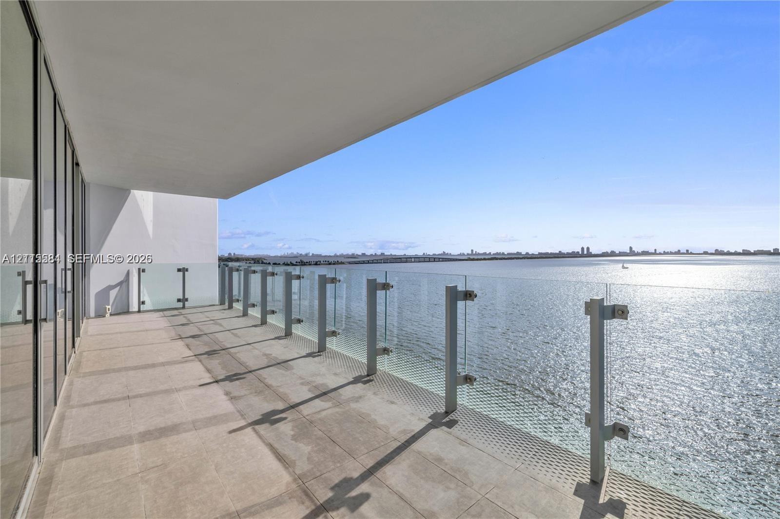 Covered balcony with large-format tile flooring and floor-to-ceiling glass wall panels, featuring a frameless glass railing with metal posts