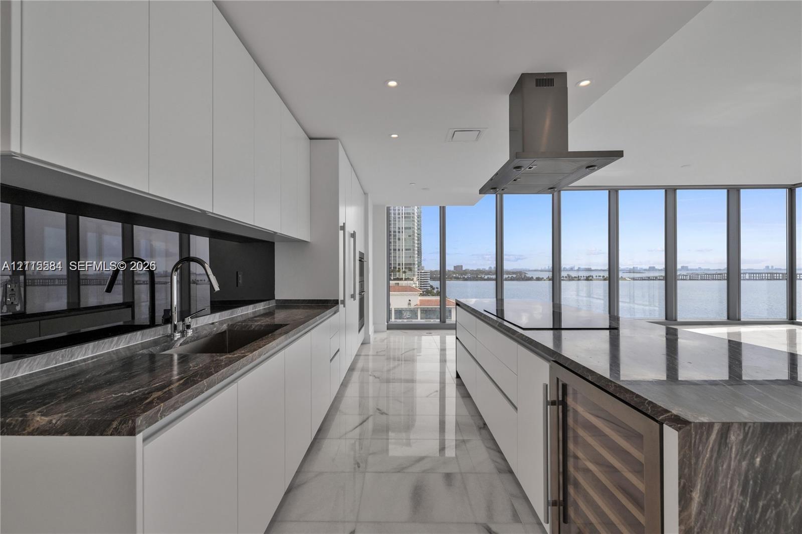 Contemporary kitchen featuring white flat-panel cabinetry, stone countertops, integrated wine refrigeration, stainless steel vent hood, and floor-to-ceiling windows