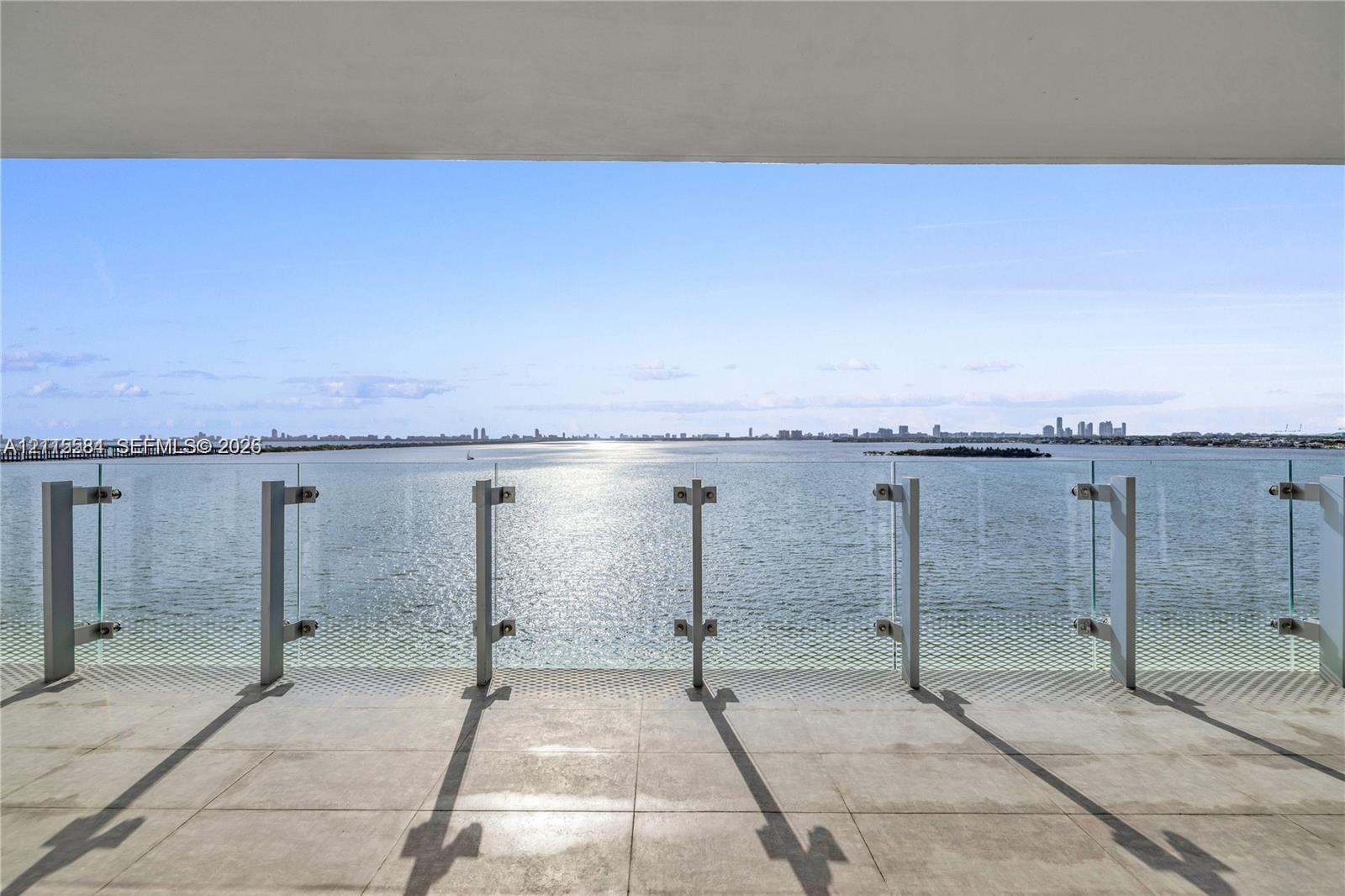 Covered terrace featuring large-format stone flooring and a glass-panel railing overlooking the water and distant cityscape