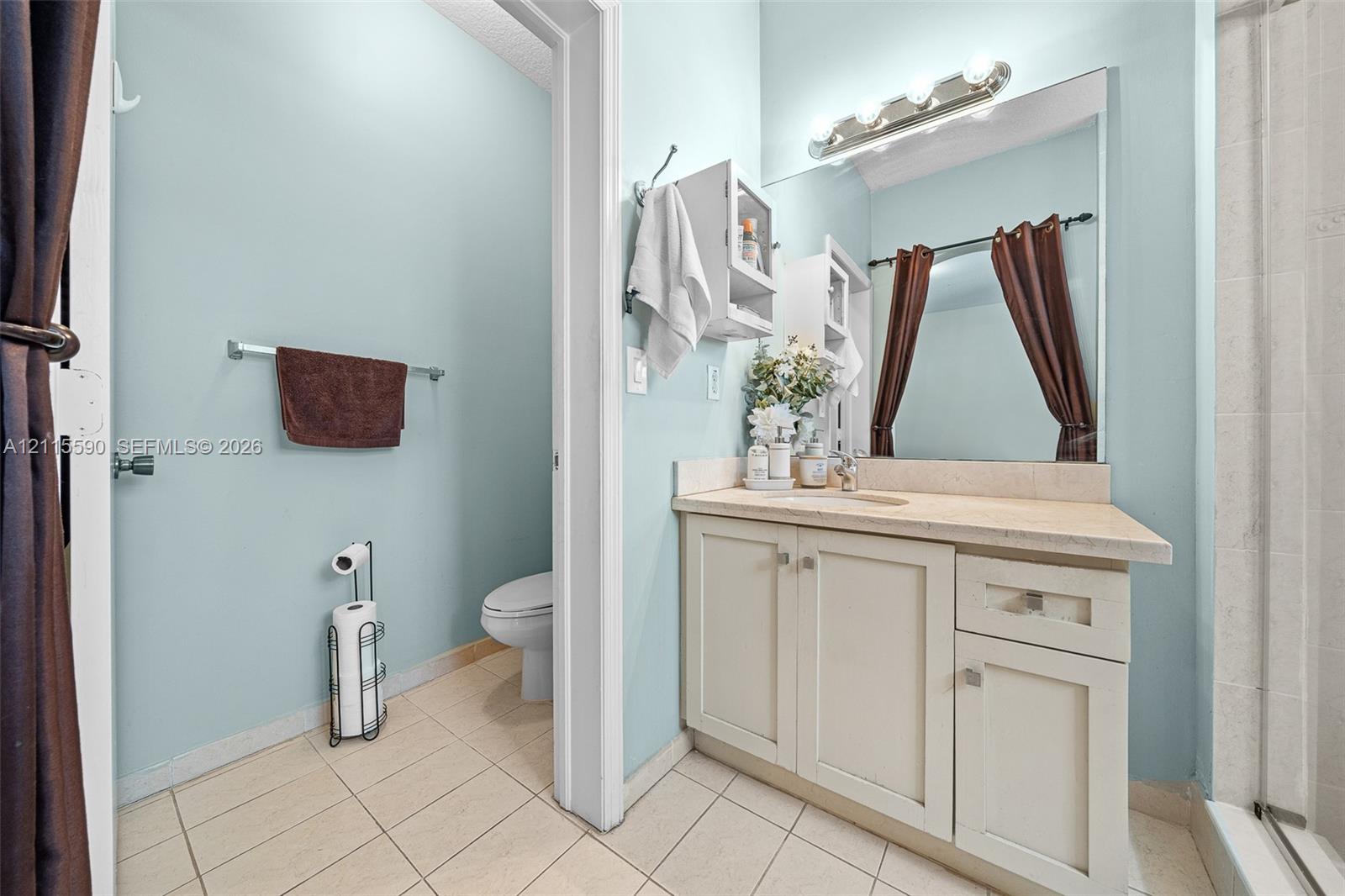Bathroom featuring a vanity with light-toned cabinetry, solid surface countertop, integrated sink, wall-mounted light fixture, and tile flooring