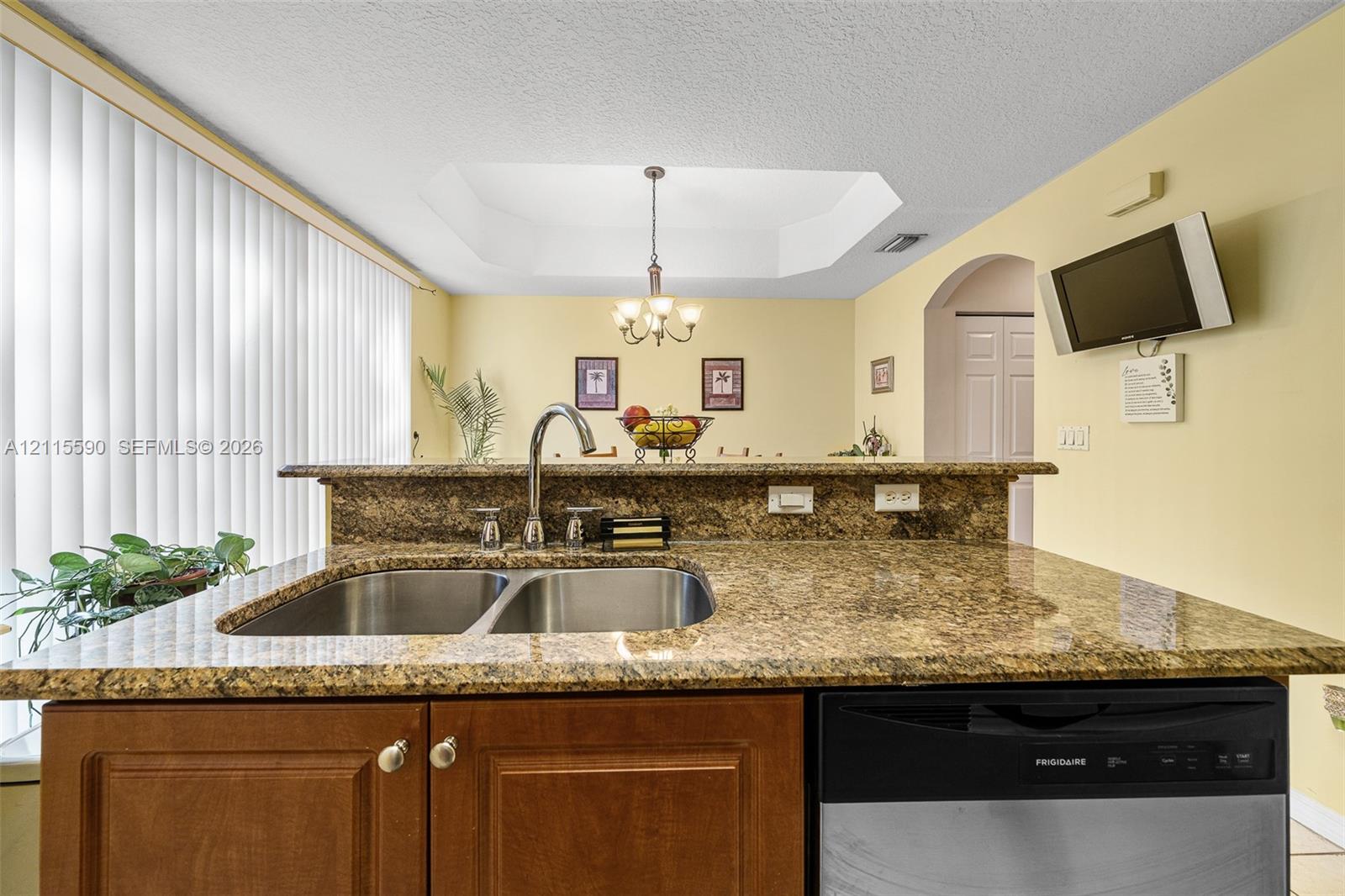 Kitchen featuring stone-finish countertops, stainless steel dual-basin undermount sink, gooseneck faucet, stained wood cabinetry, and recessed tray ceiling with a multi-light chandelier