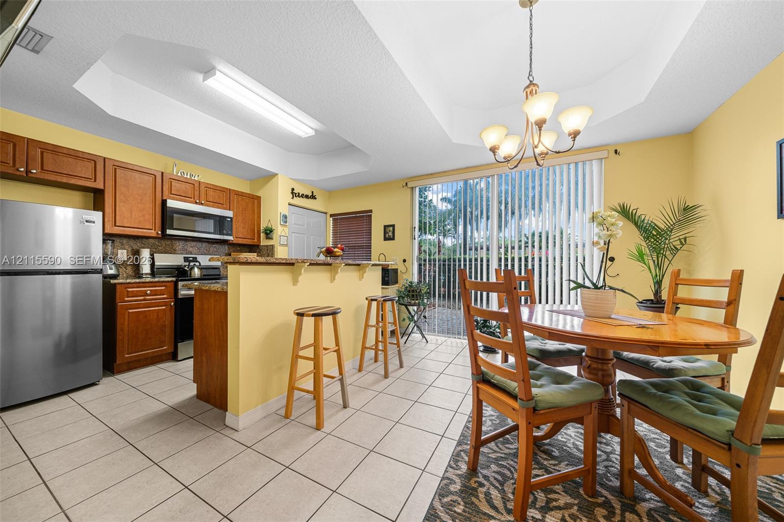 Kitchen featuring wooden cabinetry, stone-surface breakfast bar, stainless steel refrigerator, tile flooring, and a tray ceiling with a multi-arm chandelier